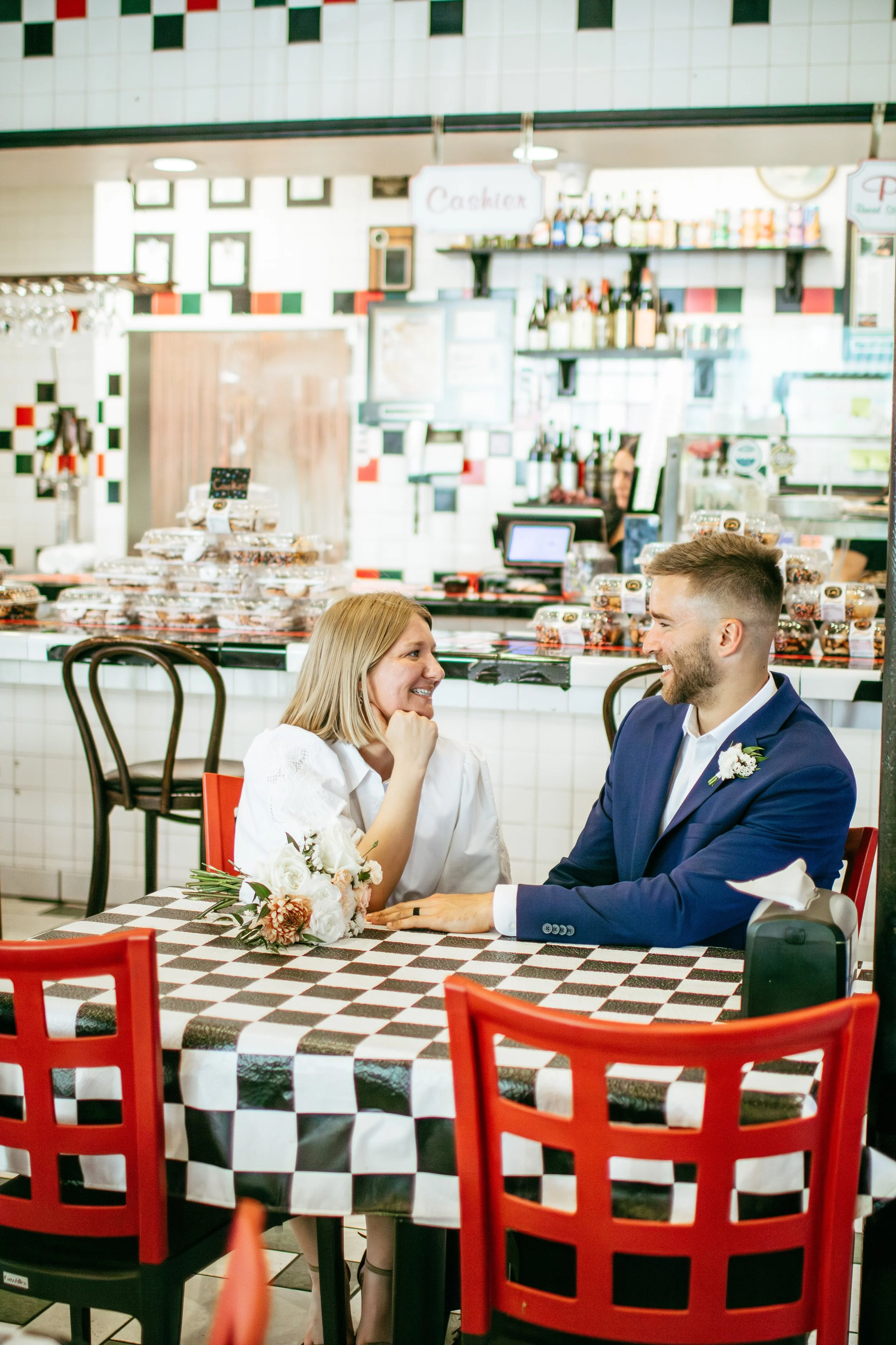 A couple sitting at a table in a restaurant, smiling and looking at each other, with one woman's hand touching her face and holding a bouquet of flowers, while the man is wearing a blue suit and a boutonniere, both with happy expressions.