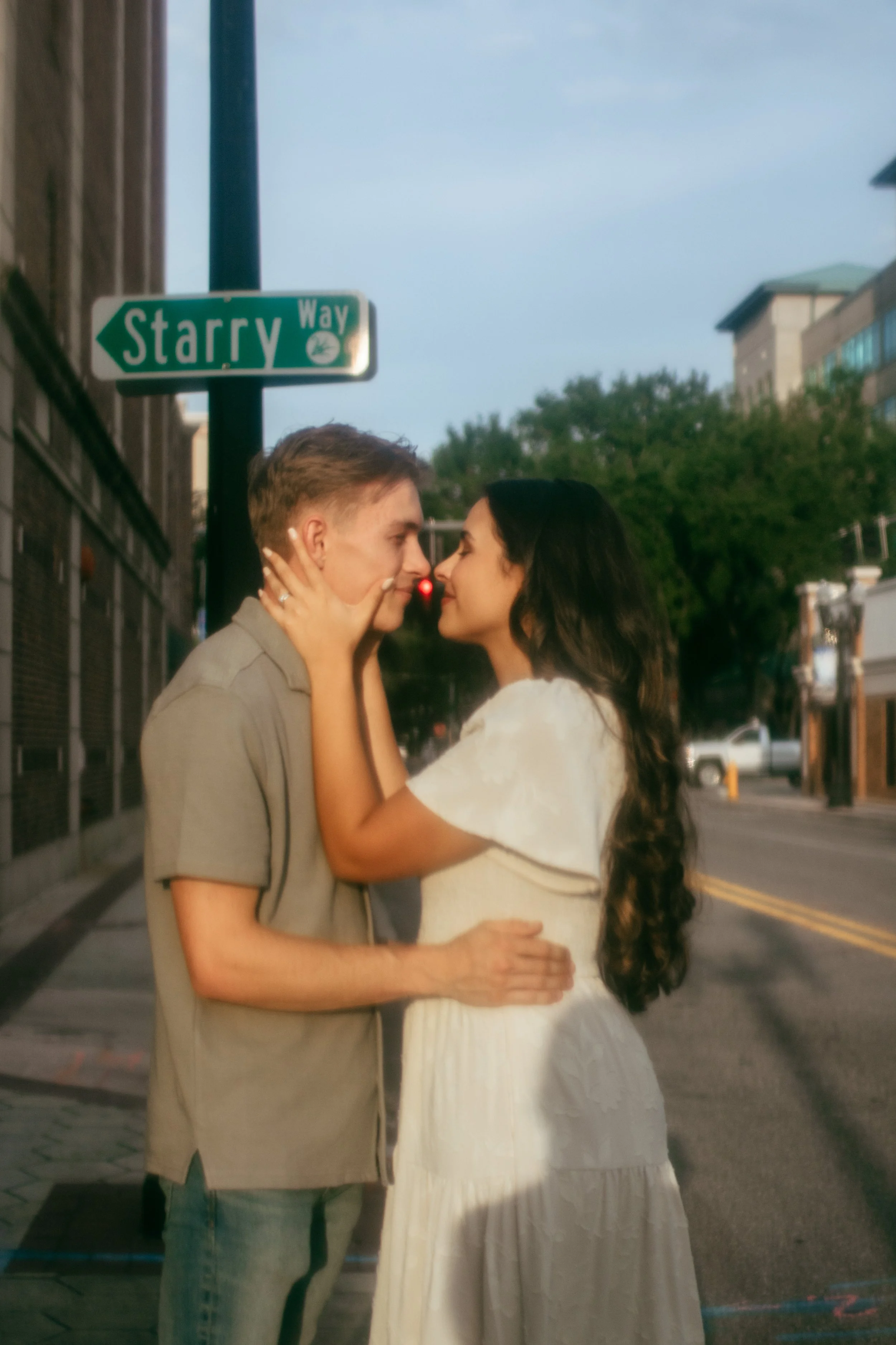 A young couple stands close together on a city street at sunset, gazing into each other's eyes, with a street sign reading "Starry Way" in the background.