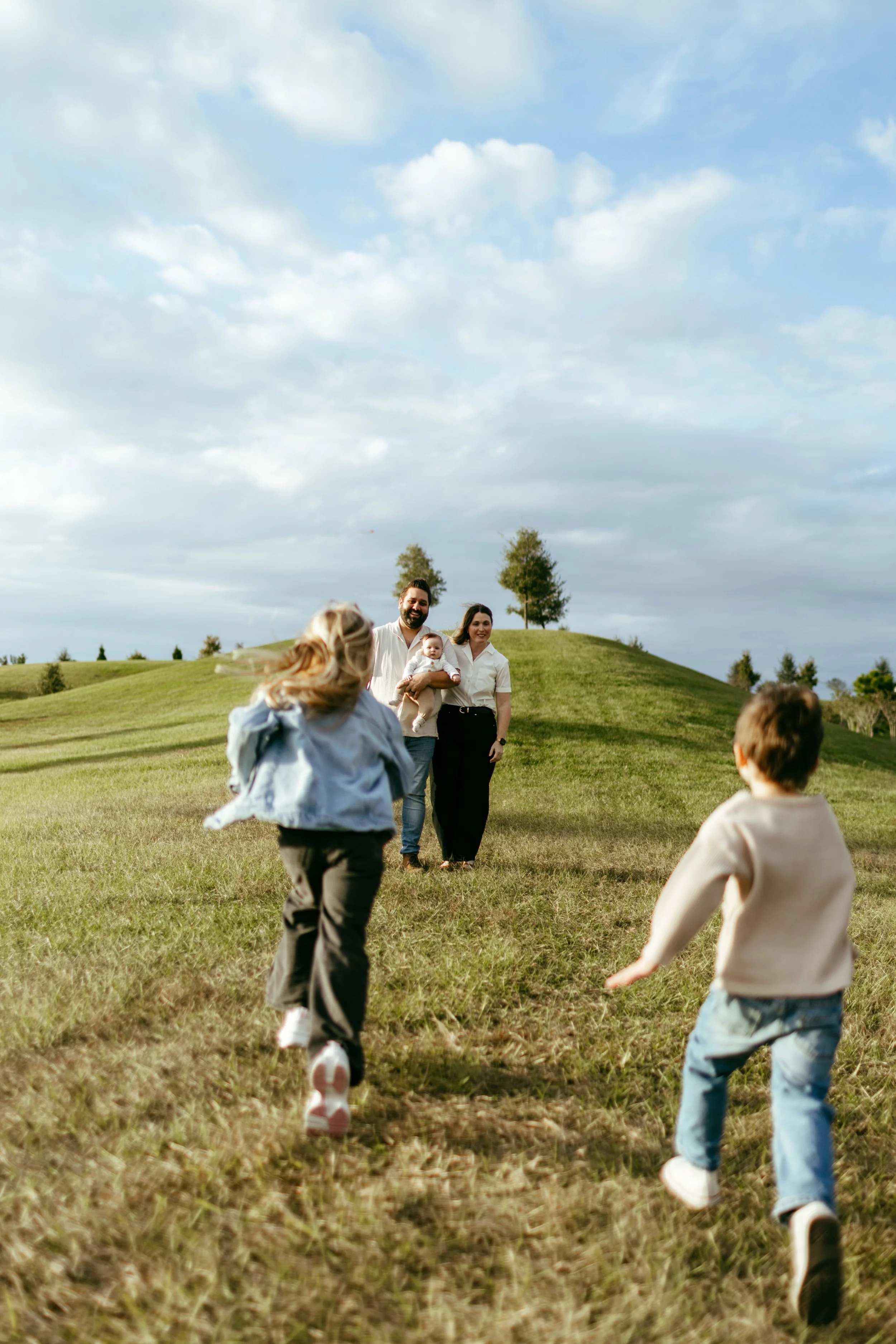 Family of four running towards parent couple holding a baby in an open grassy field under a partly cloudy sky.