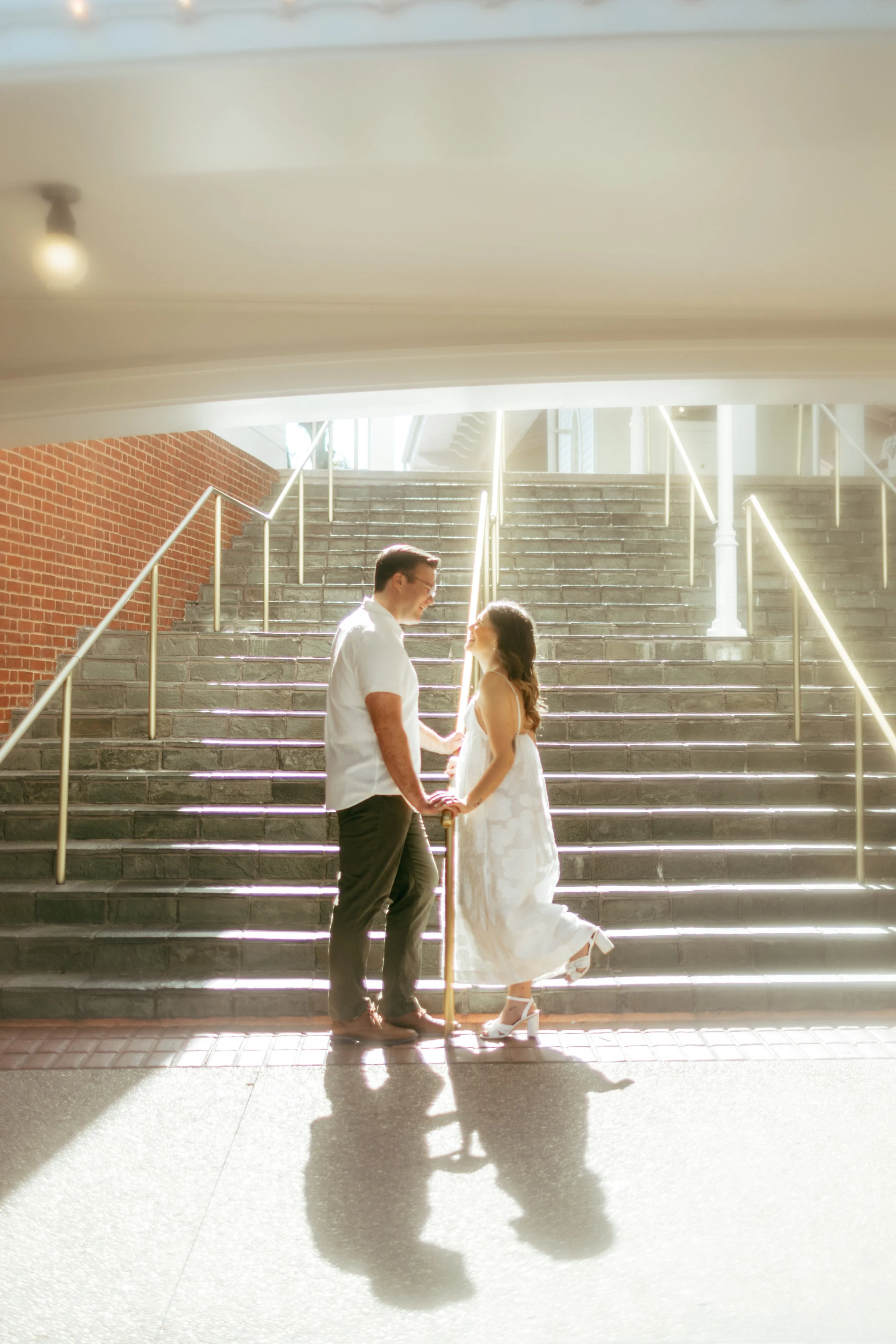 A couple holding hands and smiling at each other on stairs with sunlight shining from behind.