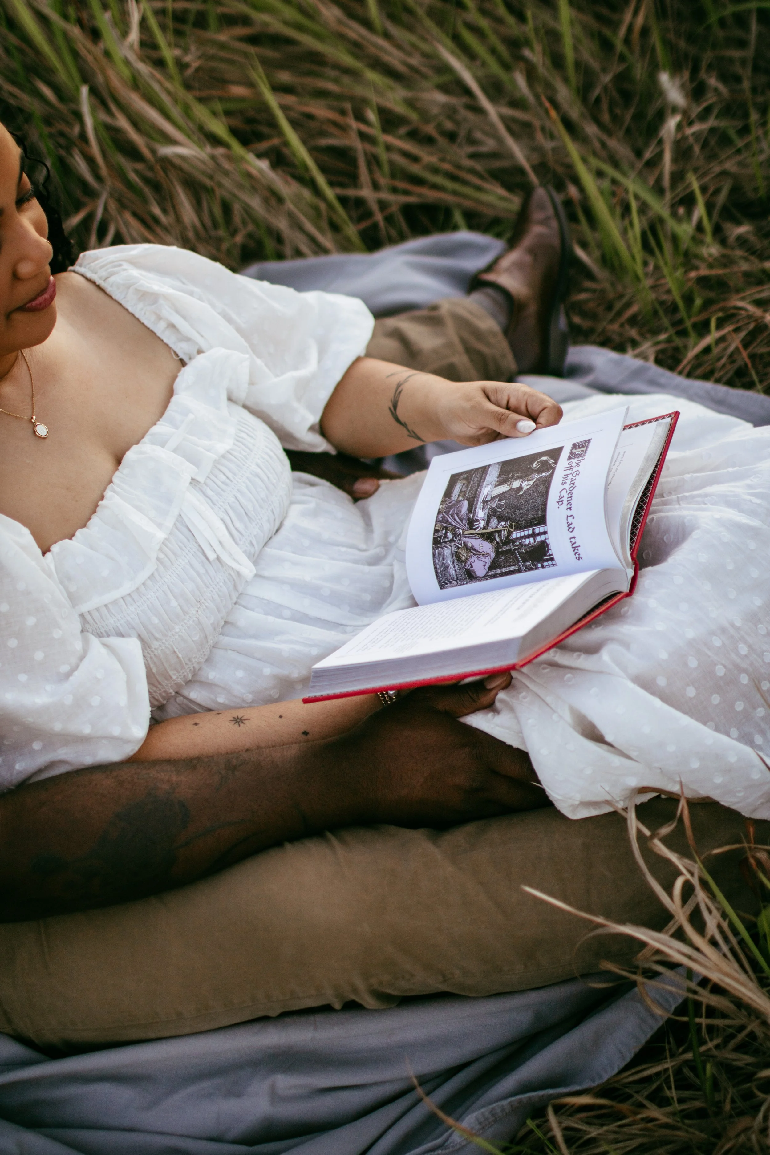 A woman in a white dress reading a book while lying on a blanket outdoors in a grassy field.