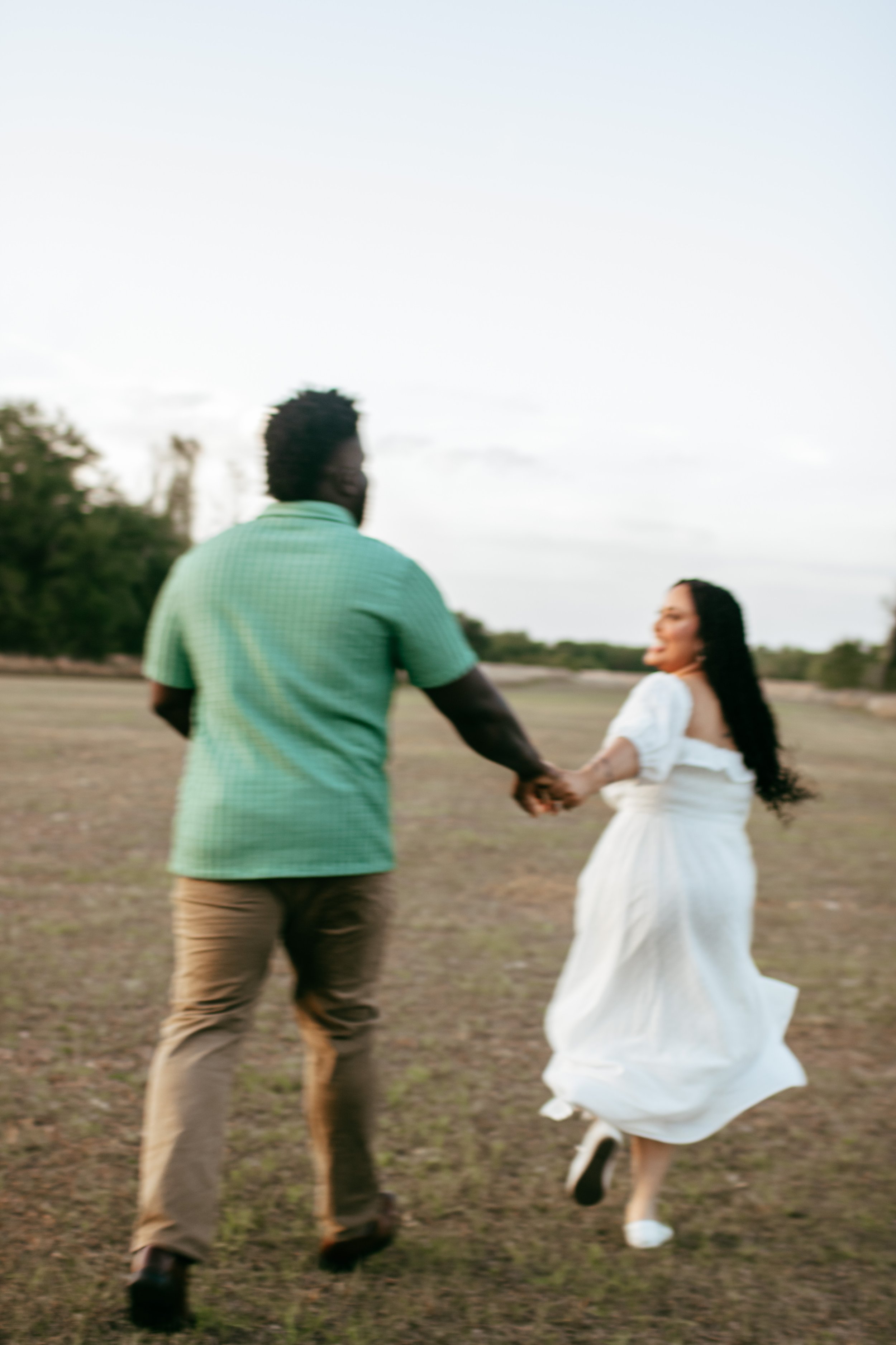 A couple holding hands and running on an open field during sunset, with trees in the background.