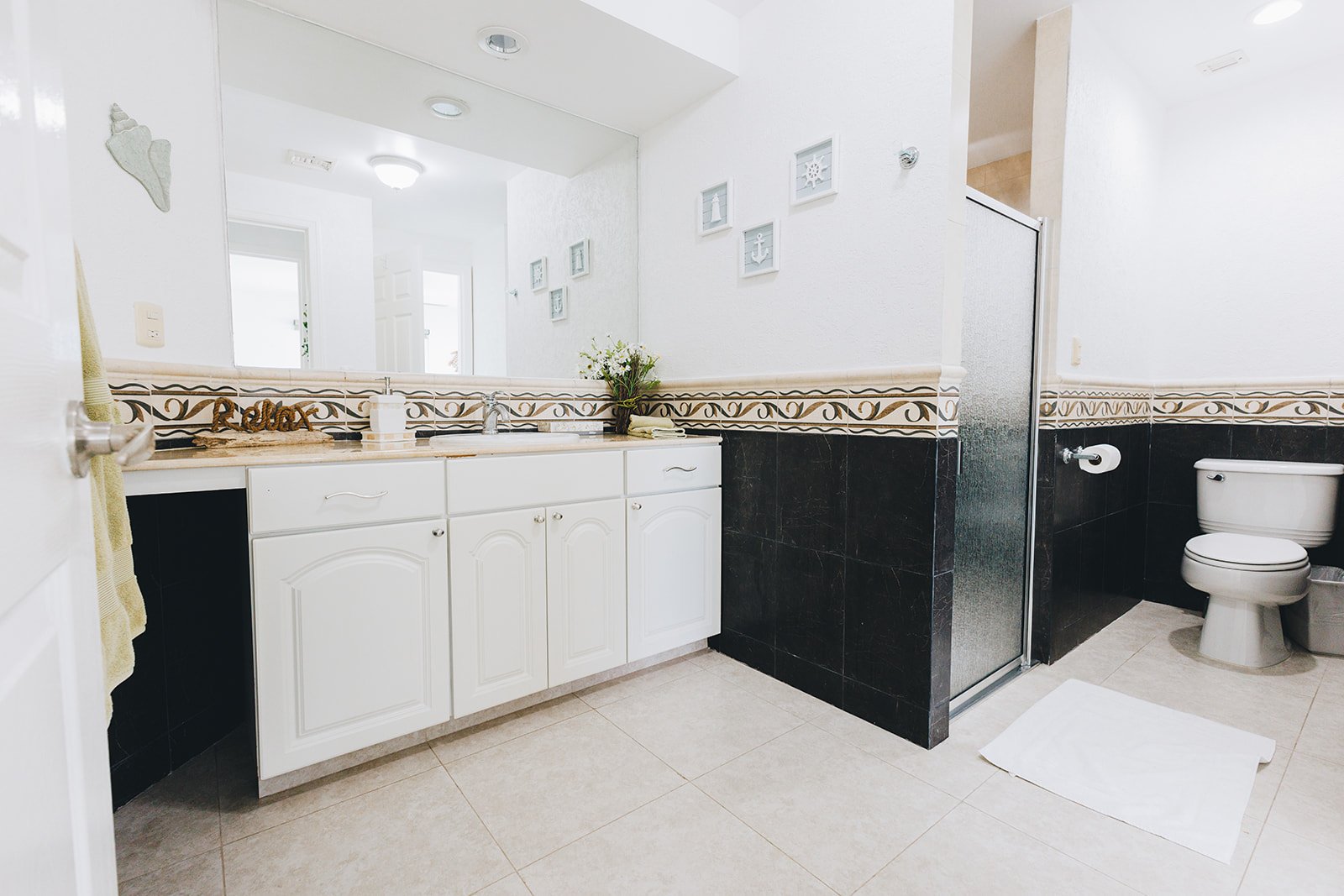 Modern bathroom with white cabinets, large mirror, and decorative wall tiles. Features a toilet, shower with glass door, and a countertop with a faucet, soap dispenser, and small plant.