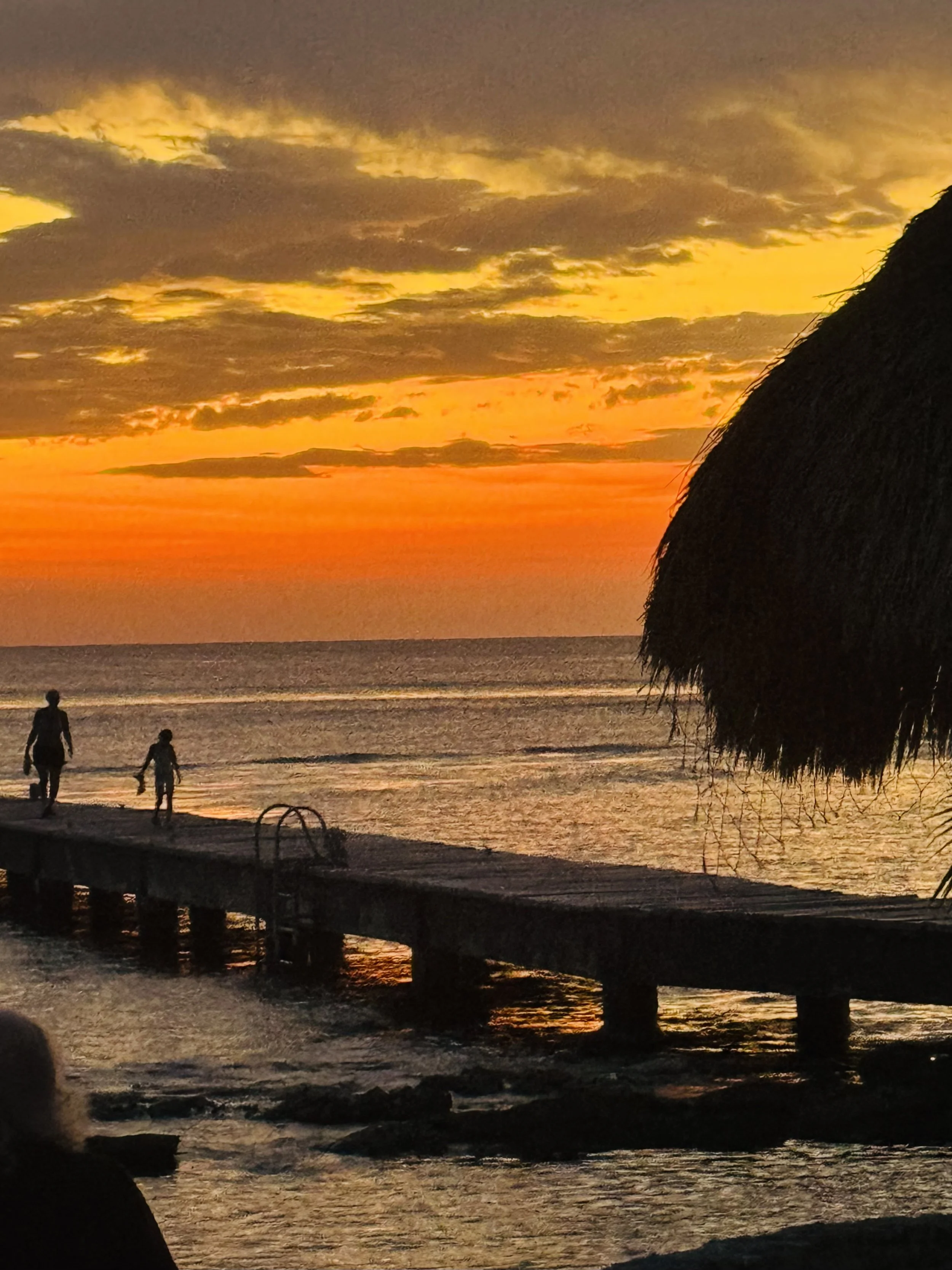 Golden Cozumel sunset viewed from We've Gone Coastal Villa — two people stroll the pier as the sky glows over the ocean, with a palapa on the right framing the perfect island evening.