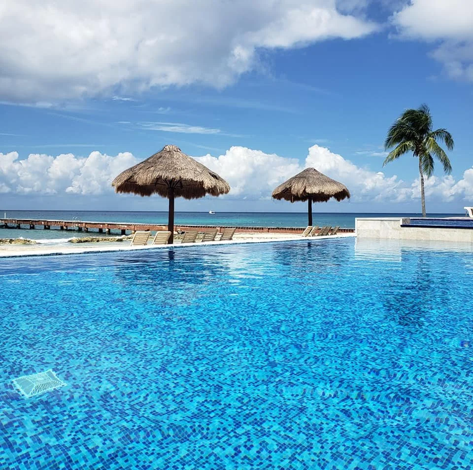 Oceanfront infinity pool at Weโve Gone Coastal Villa in Cozumel โ tropical setting with palm trees, thatched umbrellas, and stunning views of the Caribbean Sea.