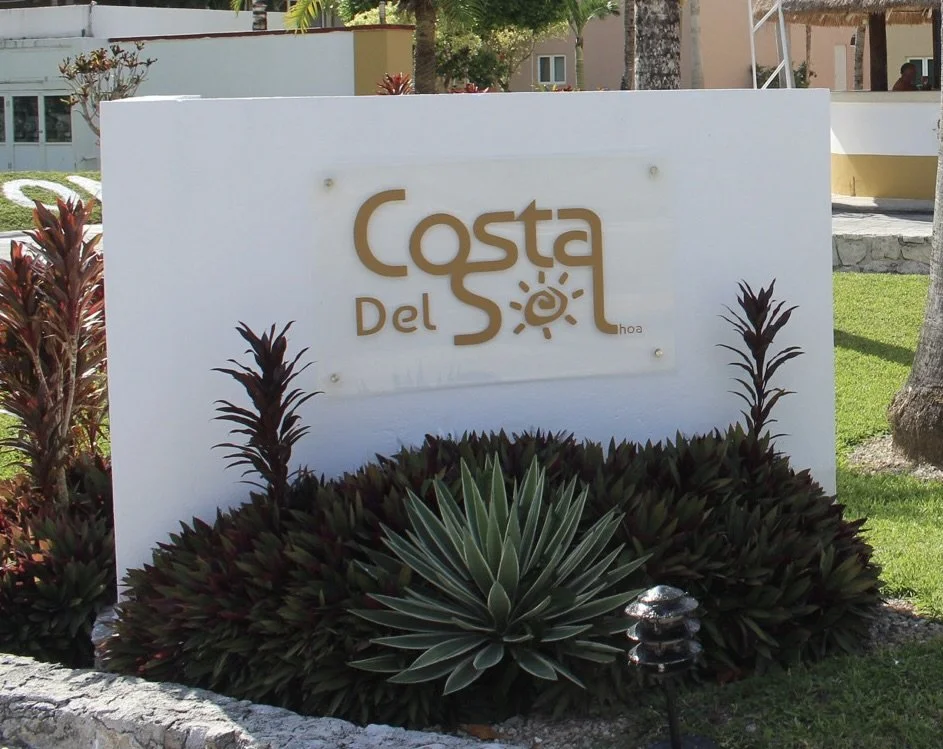 Welcome to paradise - Costa del Sol community entrance sign in Cozumel โ white sign with gold text, tropical plants, succulents, and building backdrop near Weโve Gone Coastal Villa.