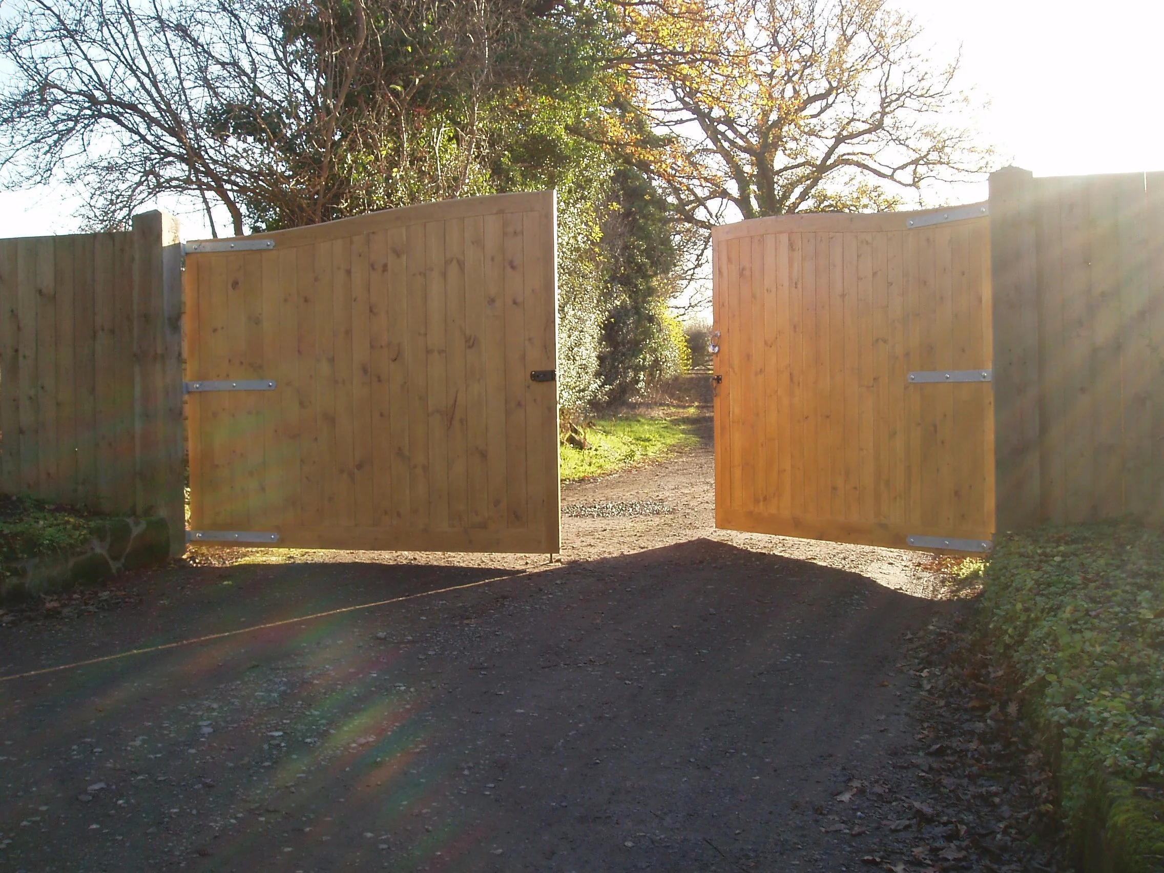 An open wooden gate on a gravel driveway, with trees and bushes in the background and sunlight shining through.