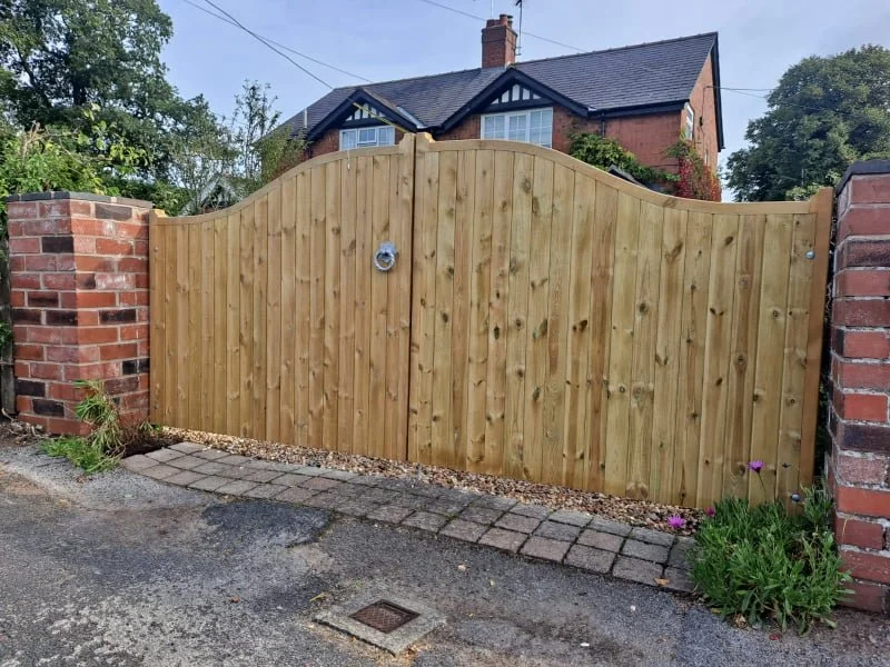 Wooden gate between brick pillars in front of a house with a slate roof and chimney, trees, and plants.