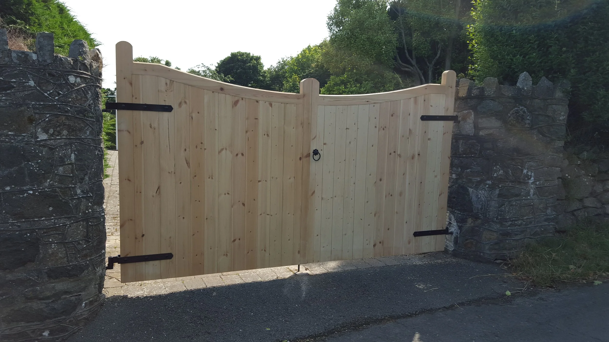 Wooden gate with curved top, attached to stone pillars on either side, situated in a garden with greenery and trees in the background.