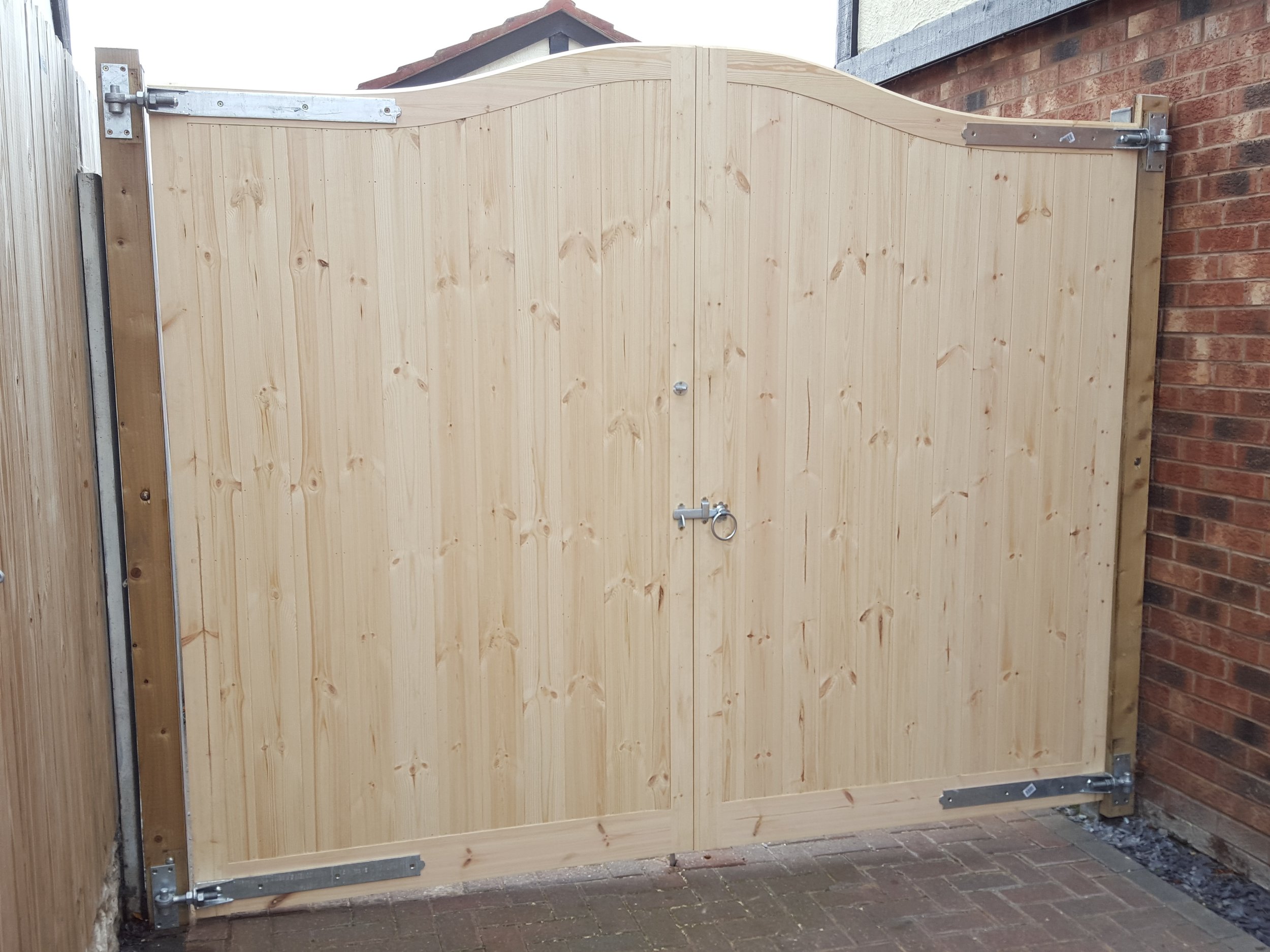 Wooden double gate with metal hinges and latch, installed between a brick wall and a wooden fence, leading to an outdoor space.