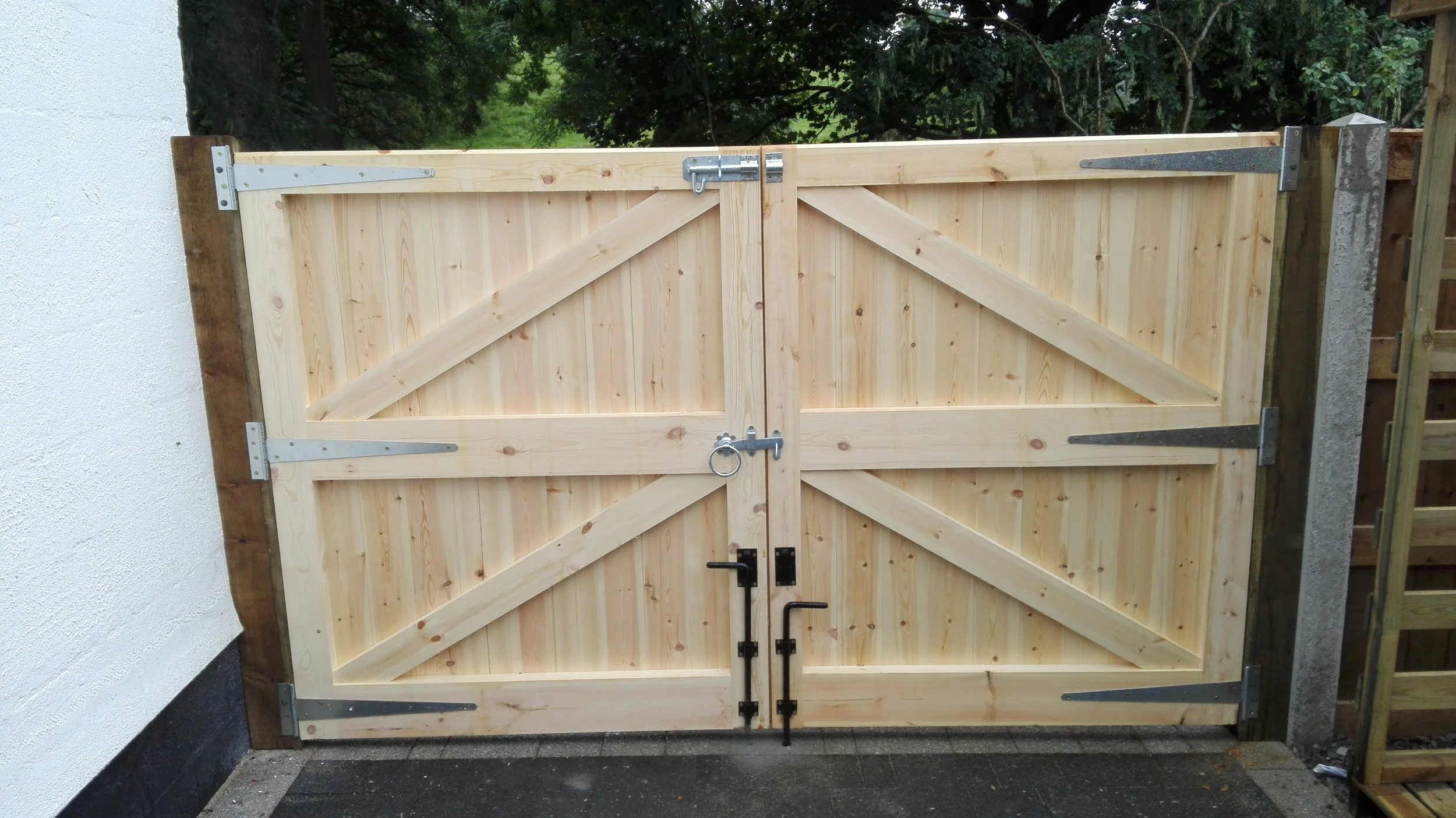 A wooden double gate with diagonal braces, secured with metal hinges, locks, and a round ring latch, situated against a white textured wall and a dark paved ground, with greenery in the background.
