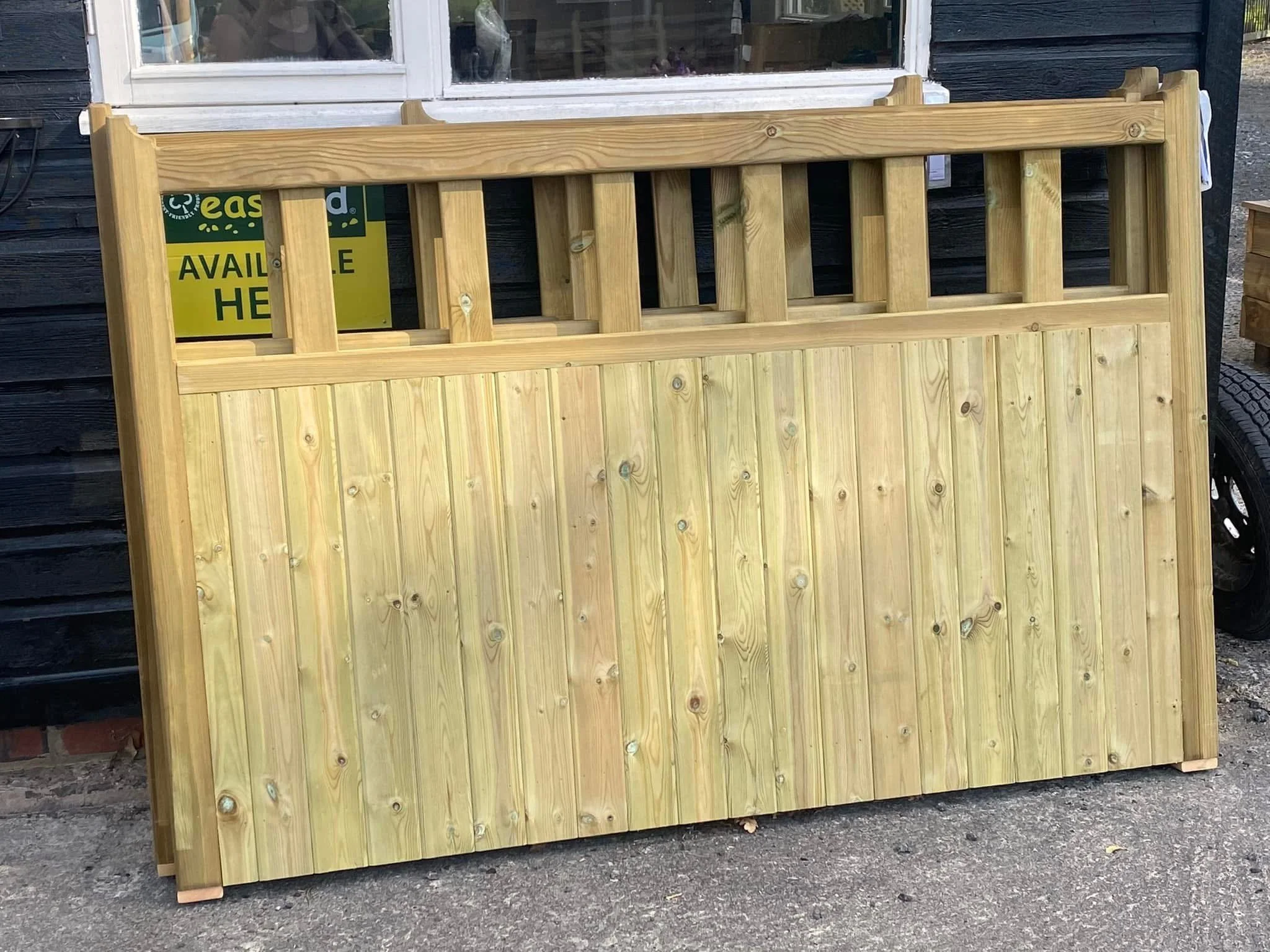 Large wooden fence panel or gate leaning against a building outside, with a window and a sign partially visible behind it.