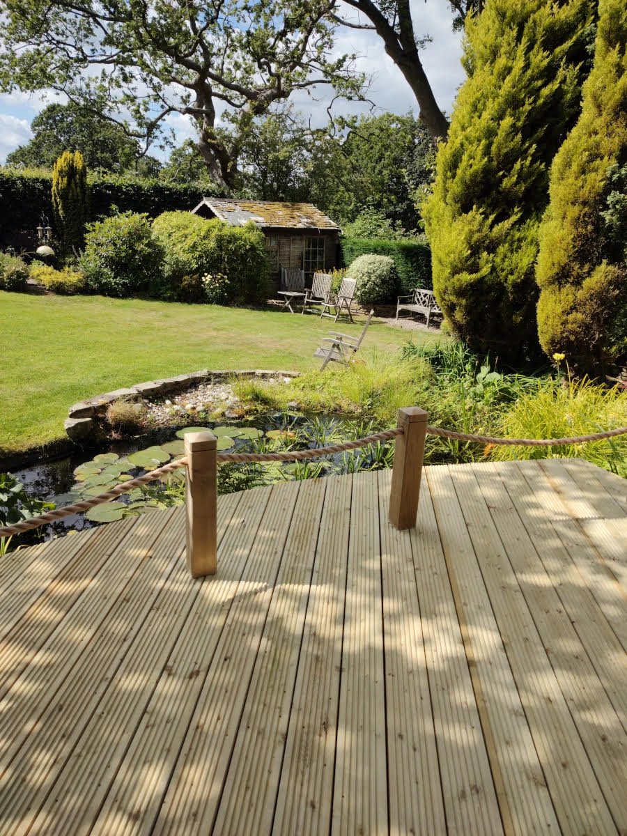 A wooden deck overlooking a lush garden with a pond, garden chairs, and a small shed, surrounded by trees and shrubs under a partly cloudy sky.