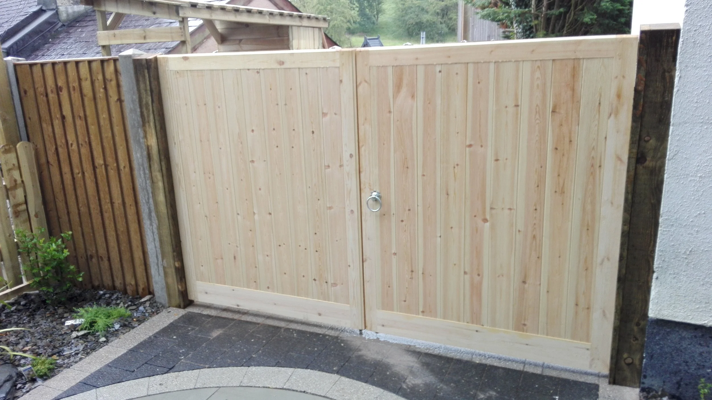 New wooden gate gate with a metal ring handle, installed between two fence posts, with a decorative stone border and garden plants nearby.