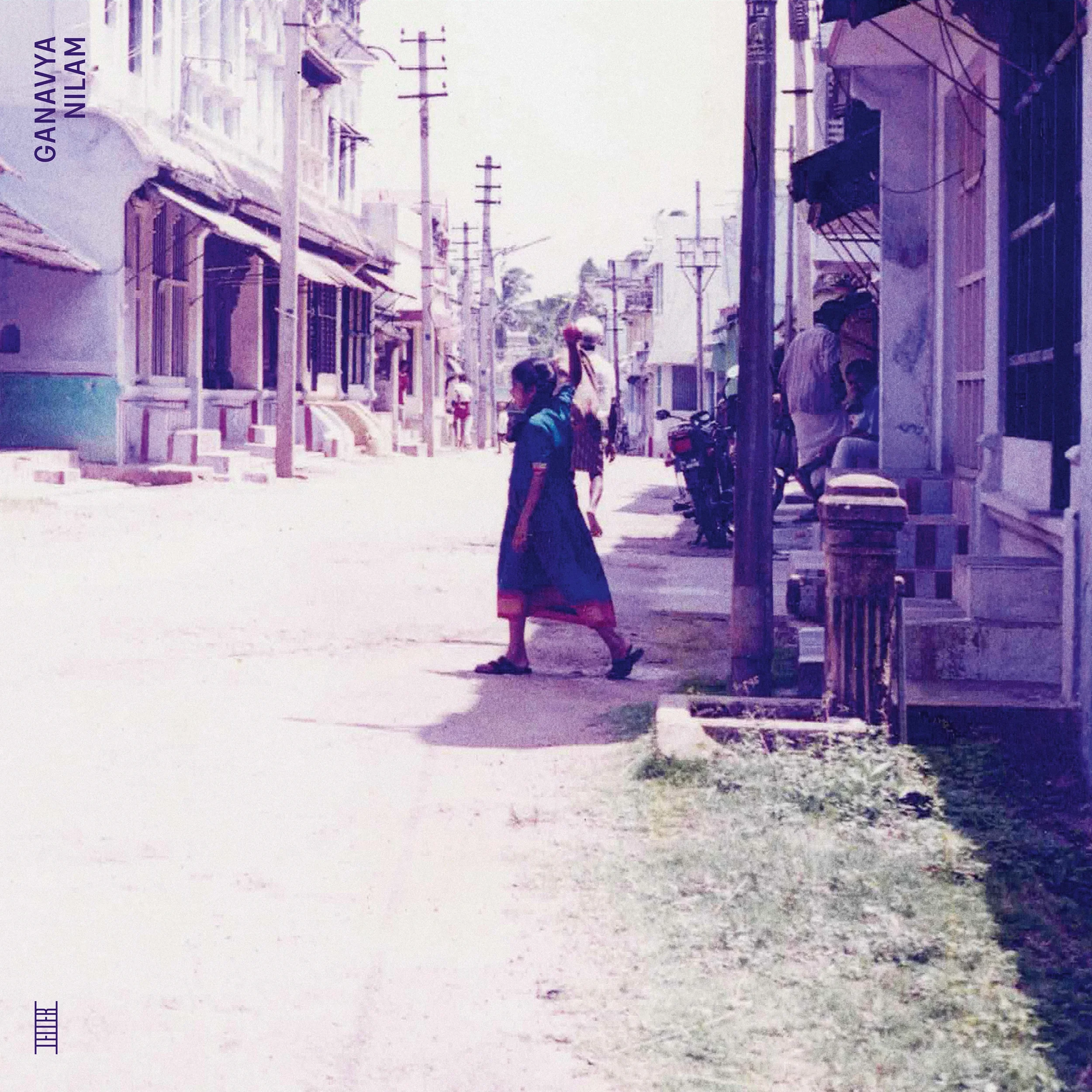 A street scene in Ganjva, Nizamabad, featuring a woman walking on the sidewalk and carrying items on her head, with other pedestrians and buildings lining the street.