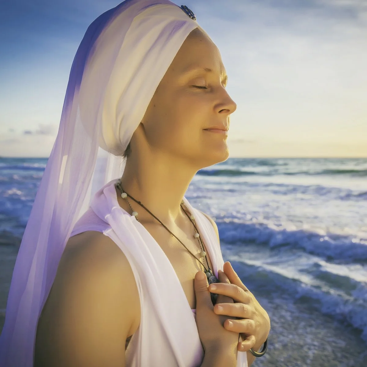 A woman with a serene expression, eyes closed, standing at the beach during sunset, holding her hand to her chest, wearing a white head wrap and jewelry.