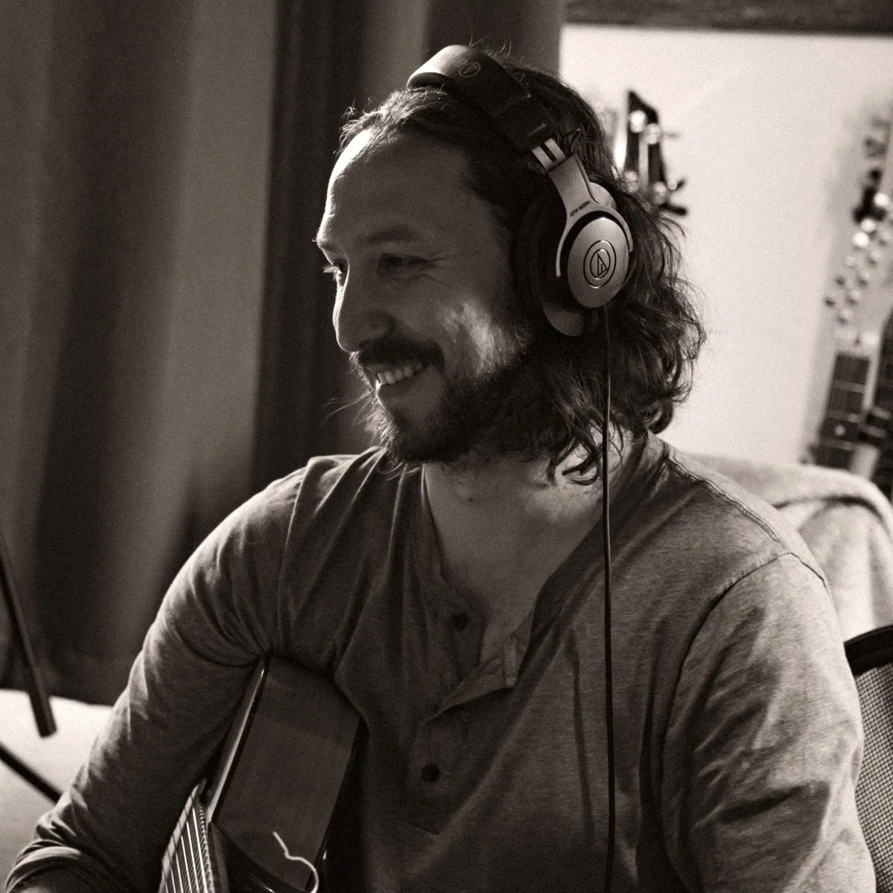 A man with long hair and a beard smiling while wearing headphones and playing an acoustic guitar.