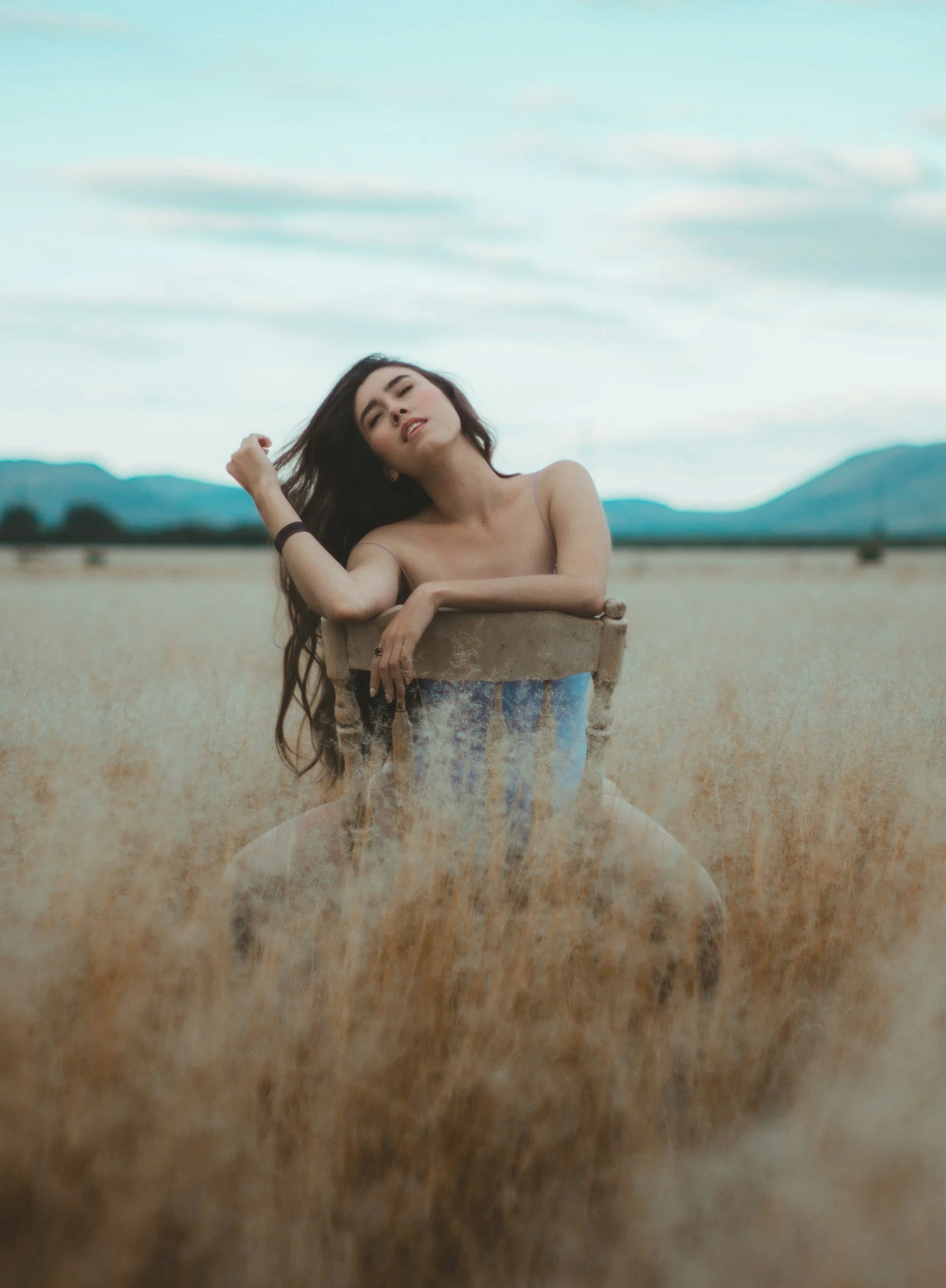 A young woman with long dark hair, reclining on a wooden chair in a vast desert landscape, with mountains in the background and a cloudy sky.