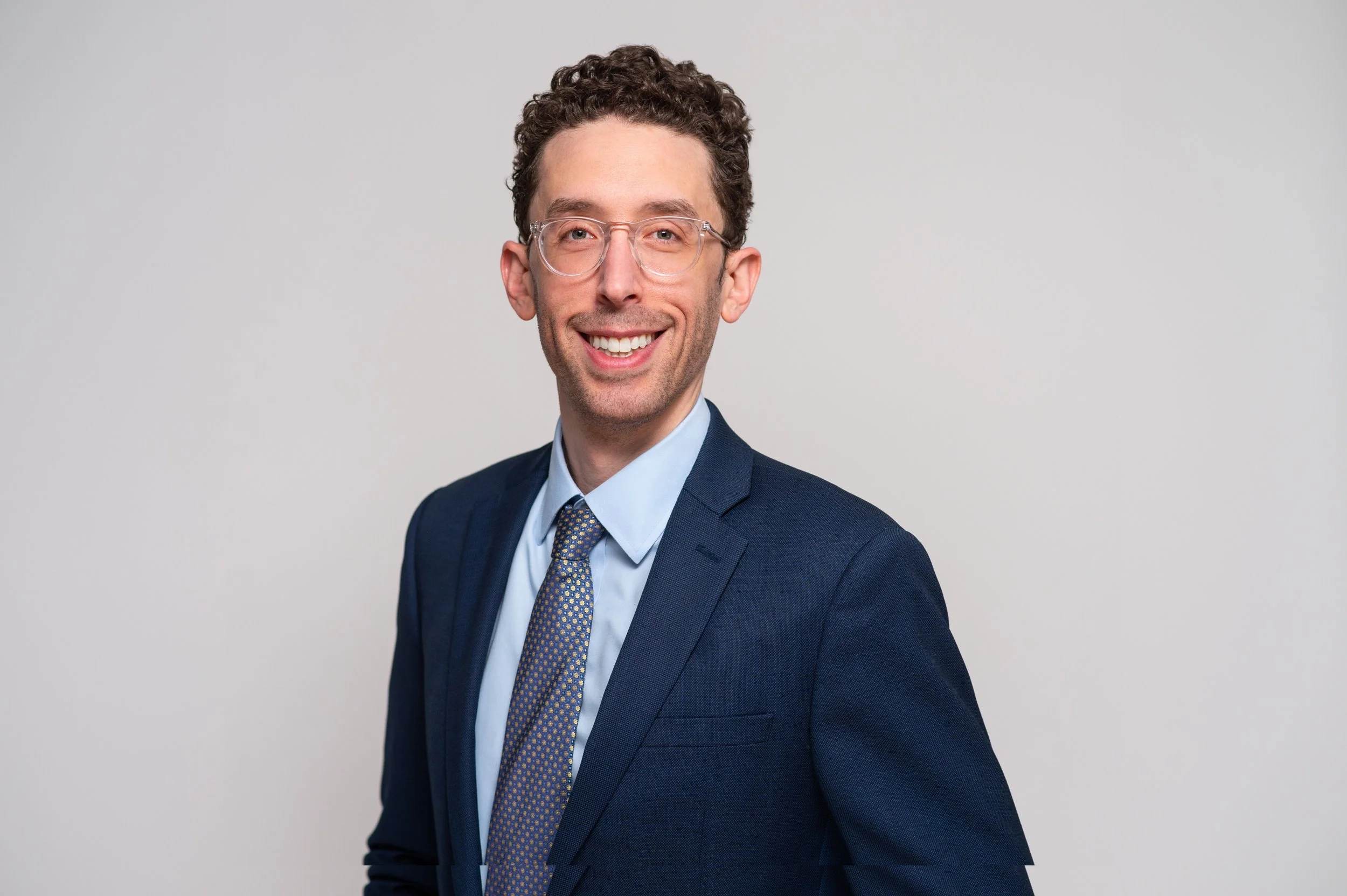 A smiling man in a navy blue suit, light blue shirt, and patterned tie, wearing clear glasses, posing against a plain light grey background.