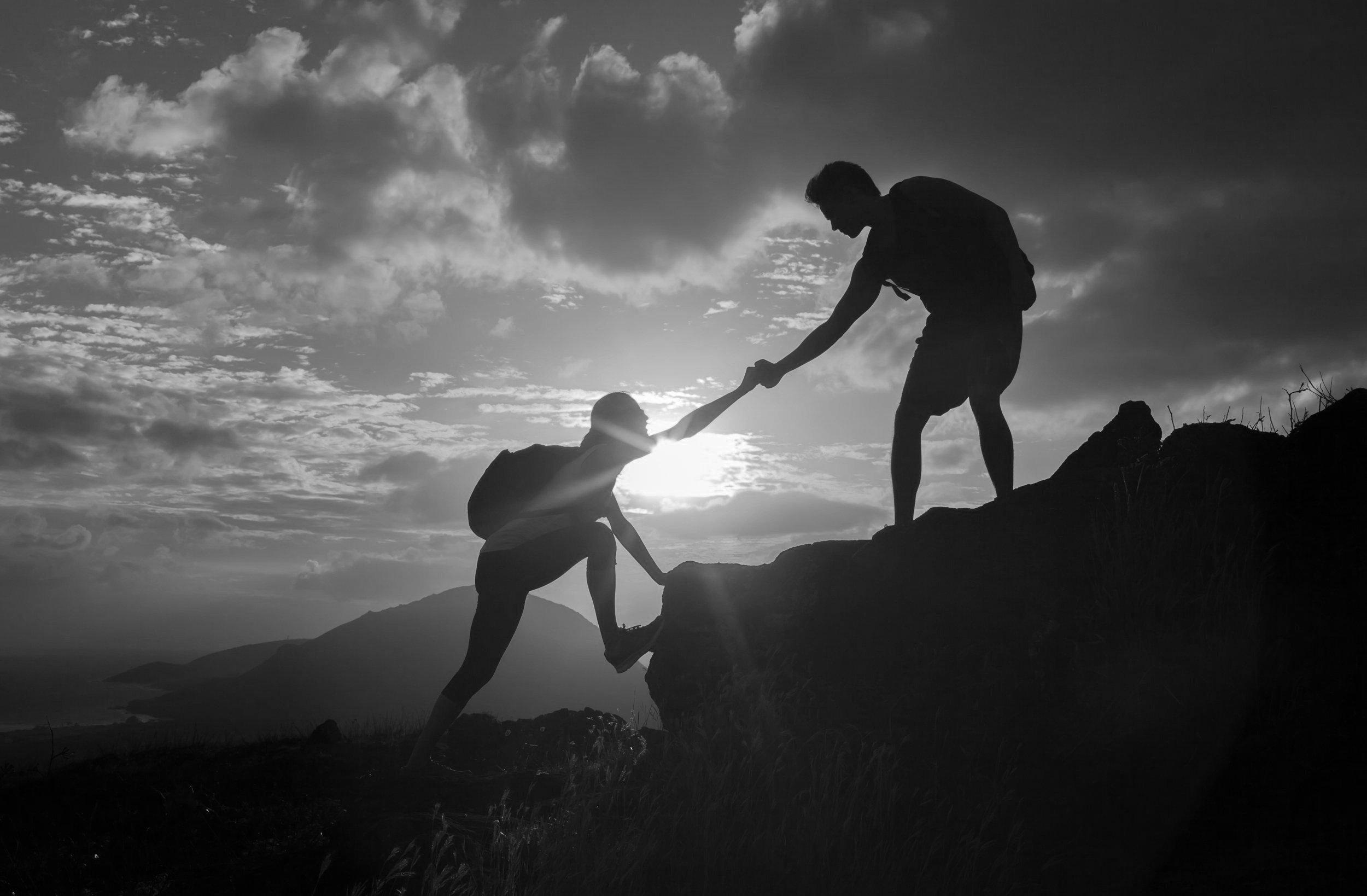 Silhouette of two hikers helping each other climb a mountain at sunset.