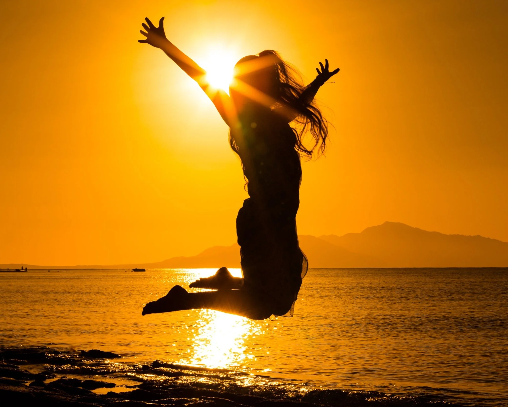 Silhouette of a person jumping on a beach at sunset with sunburst effect, ocean, and mountains in the background.