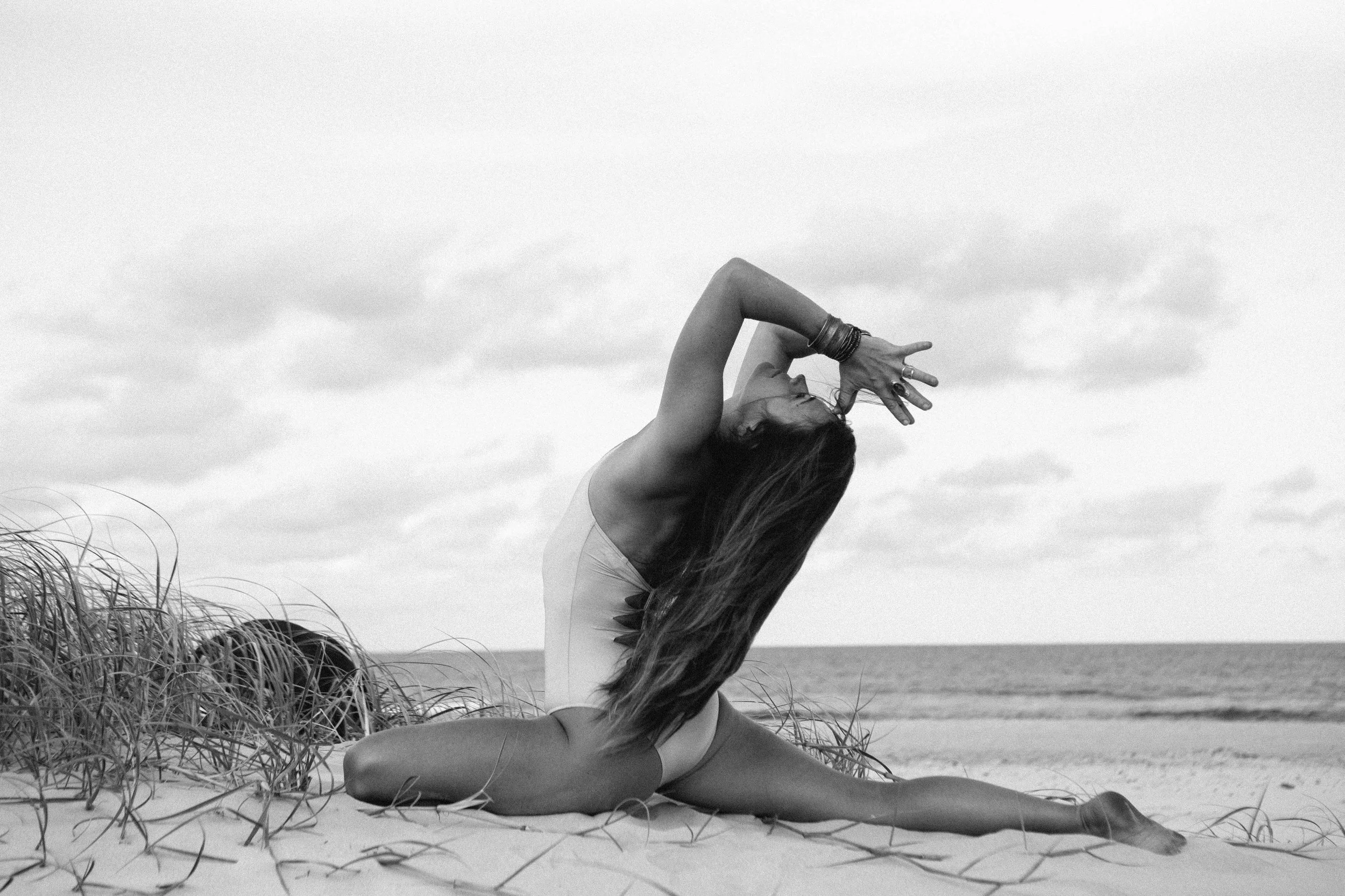 A woman in a yoga pose on a sandy beach with grass, ocean, and cloudy sky in the background