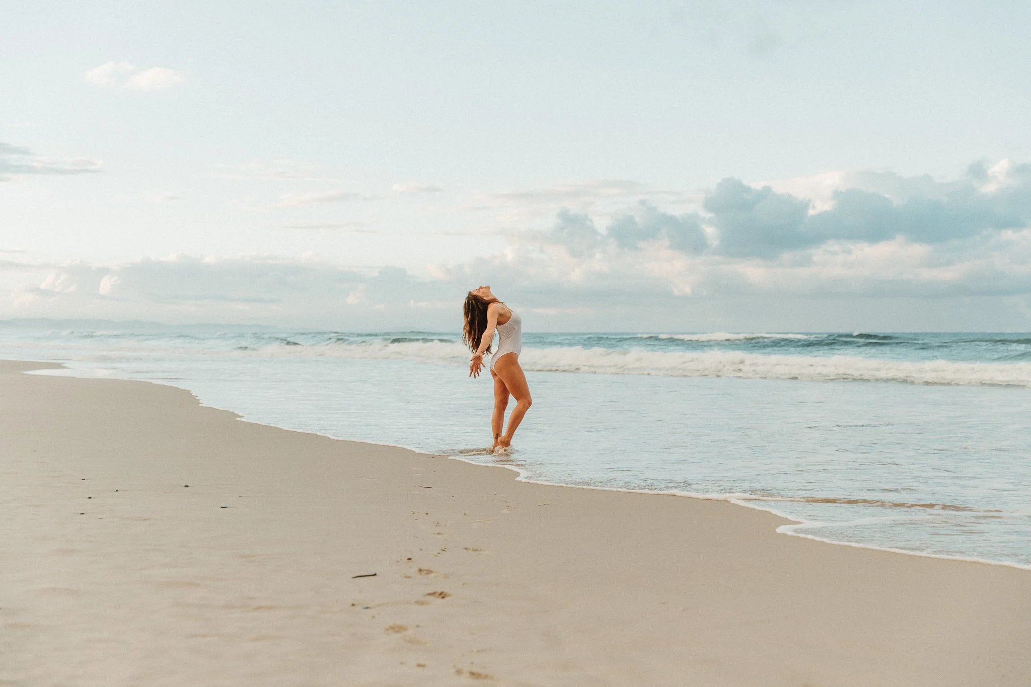 A woman with long hair in a white swimsuit standing at the shoreline on a sandy beach, with waves in the background and her head tilted back.