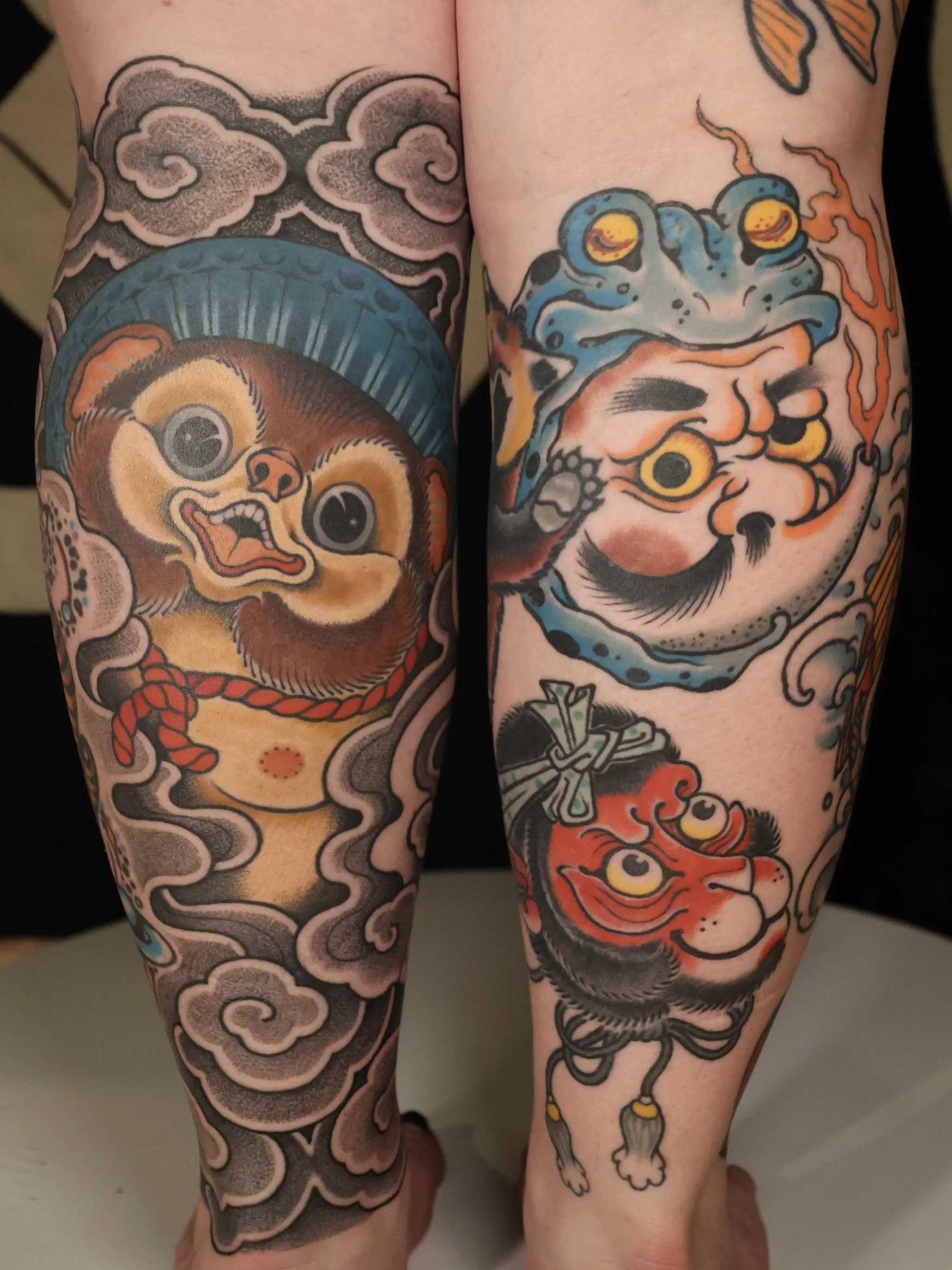 Japanese Leg Tattoos of Tanuki, Hyottoko , saru mask in bold colors and black / grey storm clouds.