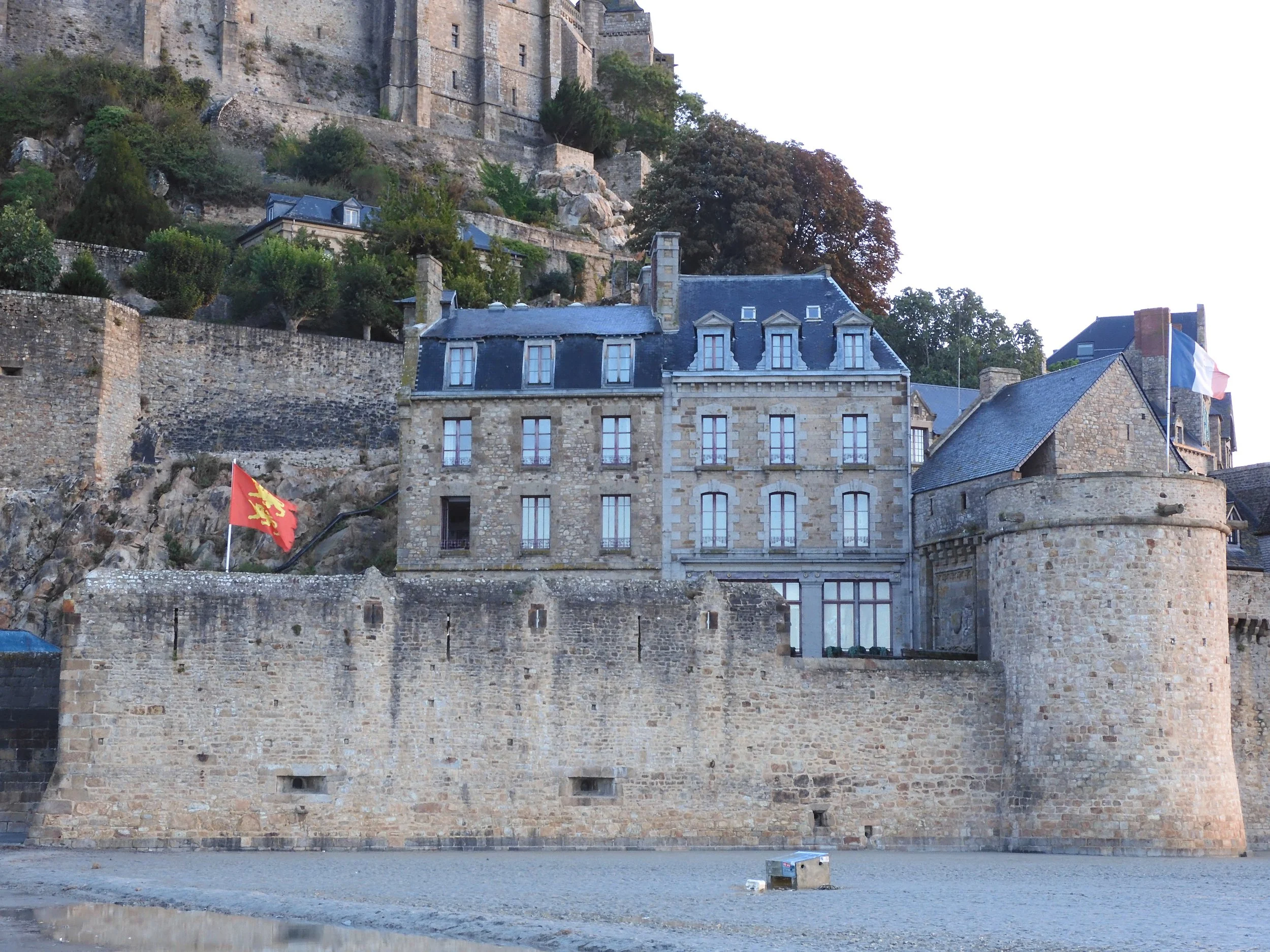 Fortified walls and defensive tower of Mont Saint-Michel rising above the tidal flats, with the red Norman lions flag flying above the ramparts.
