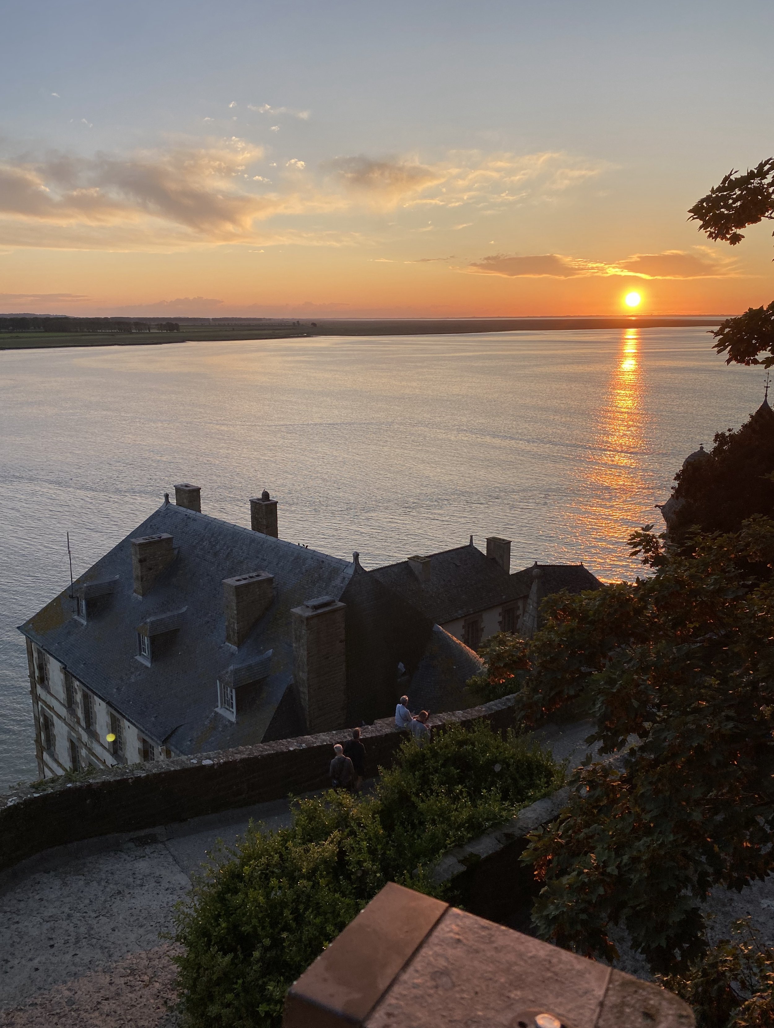 Sunset over the bay of Mont Saint-Michel viewed from the island walls, with rooftops and the golden reflection of the sun across the water.