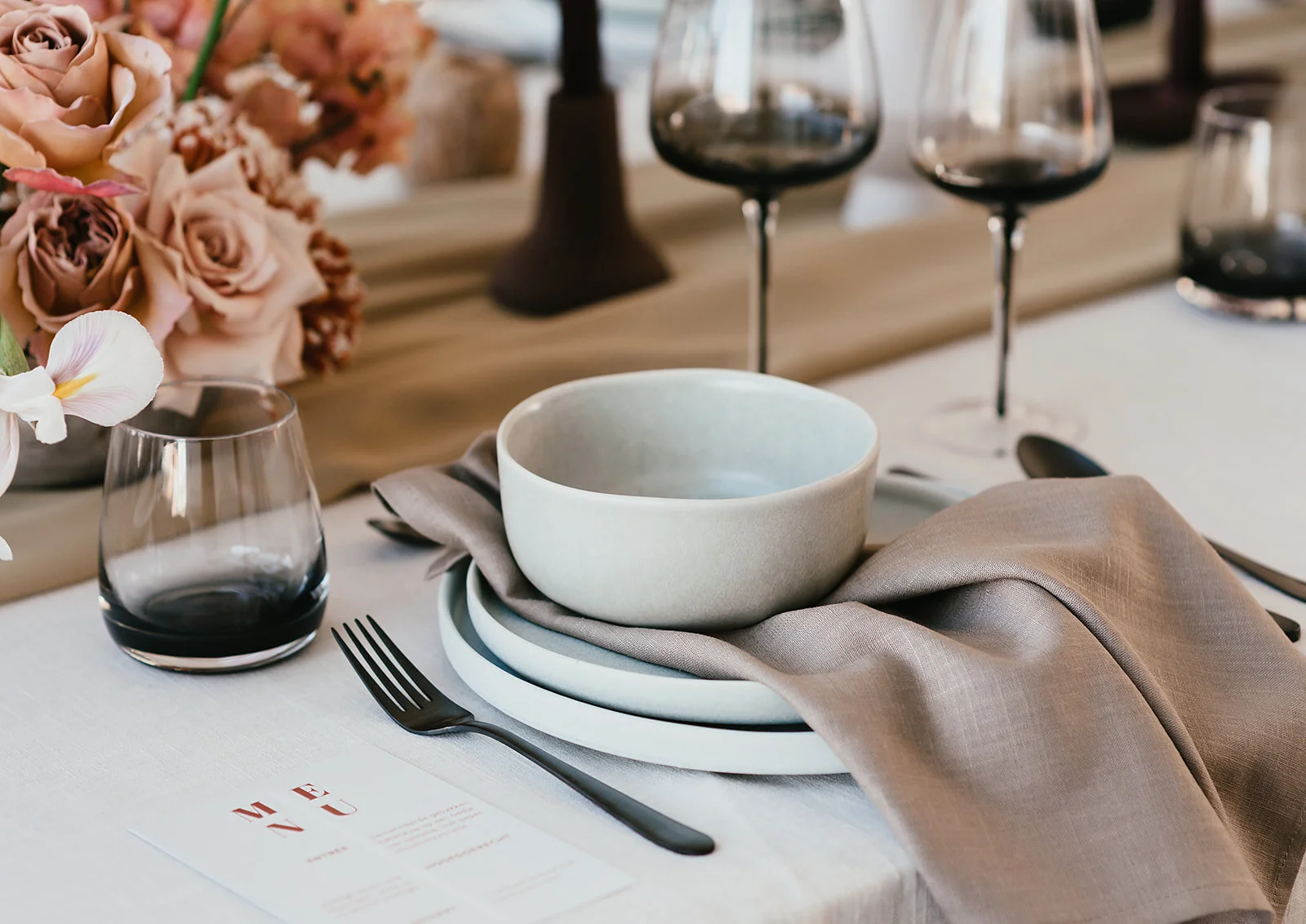 Elegante tafelstyling met lichtgrijs keramiek (een kom, dessertbord en dinerbord), een taupekleurig servet is gedrapeerd over het bord. Over het midden van de tafel ligt een loper van chifon. Het bestek en het glaswerk is zwart.