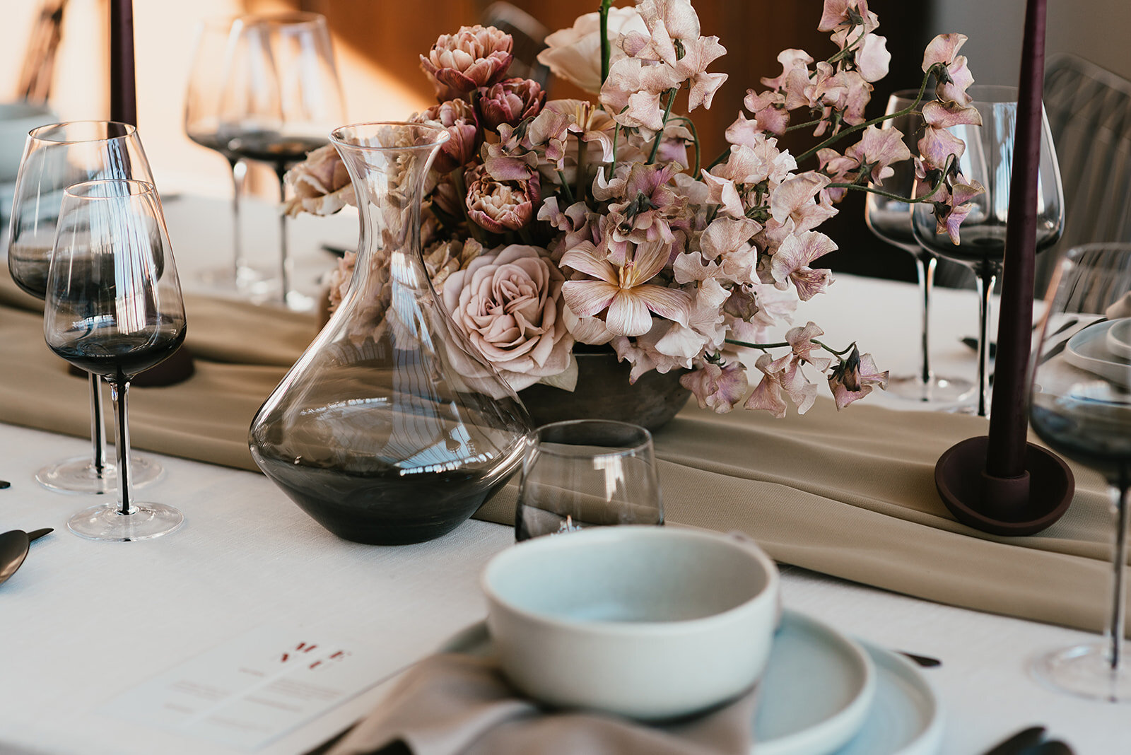 Tijdloze en stoere tafelstyling met donkere details. Zakelijke kleuren, niet uitbundig. Licht 3-delig servies ligt op tafel. Er staat zwart smoke glaswerk: wijnglazen en een decanter. Het lichtroze bloemstuk past naadloos in de setting. Lange kaarsen