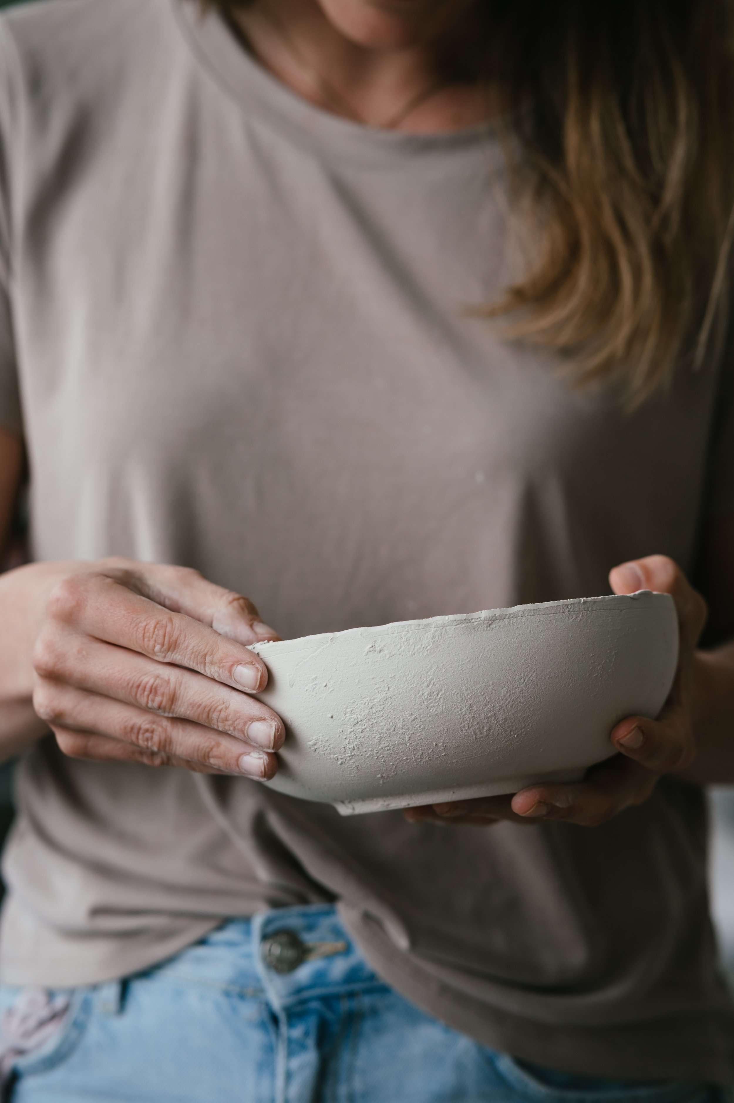 Person holding a clay bowl, wearing a beige shirt and jeans.