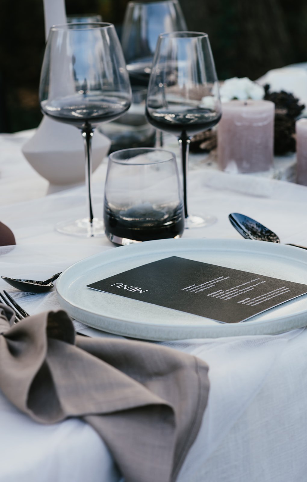 Elegant table setting with wine glasses, a plate, a menu card, cutlery, and candles on a white tablecloth.