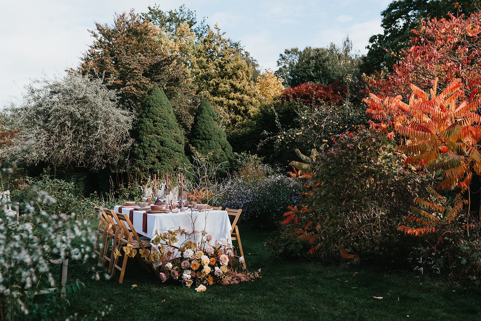 Outdoor dining table setup amidst autumn foliage with chairs and floral decorations.