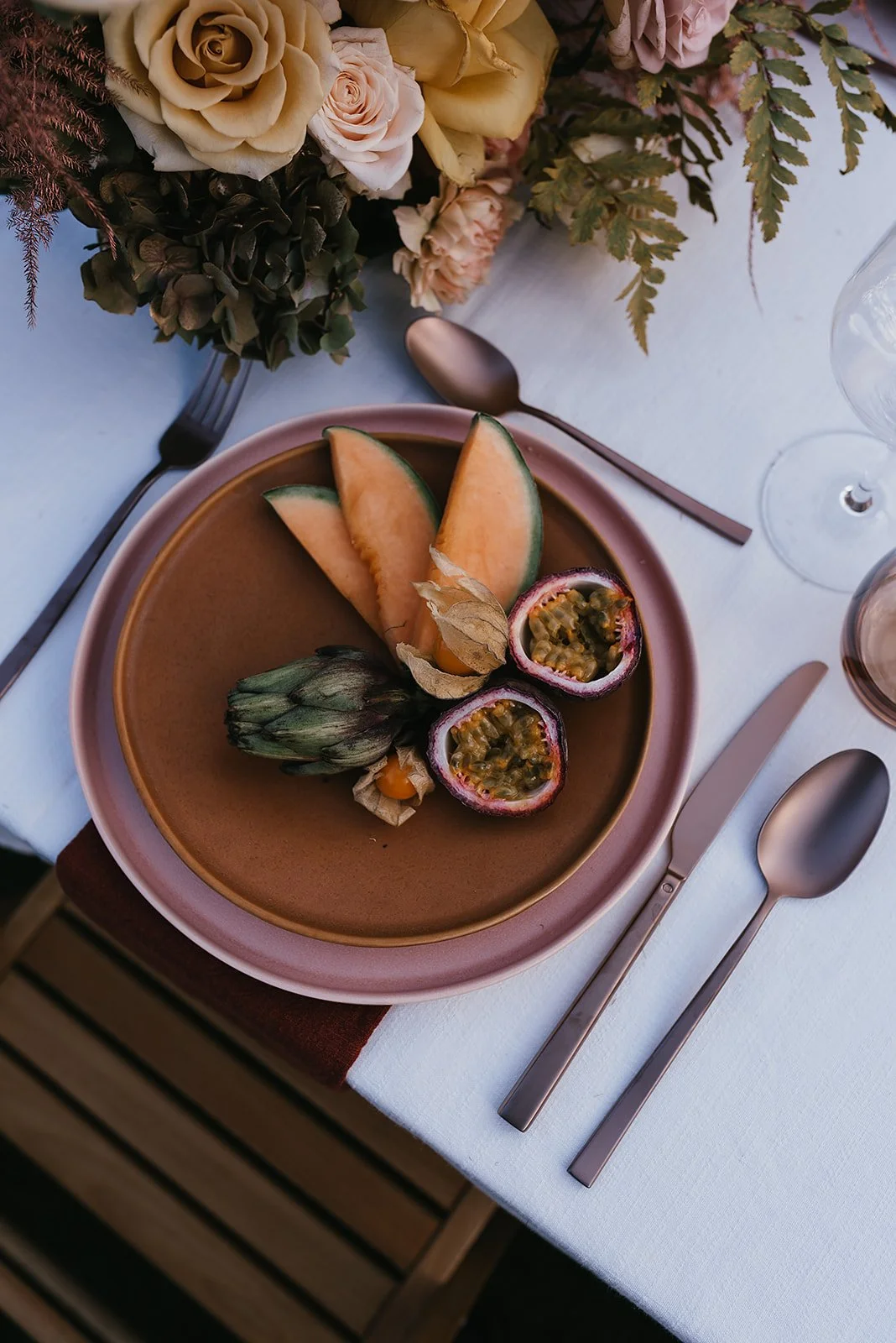 Plate with sliced cantaloupe, passion fruit, artichoke, and physalis, surrounded by cutlery and floral arrangement.
