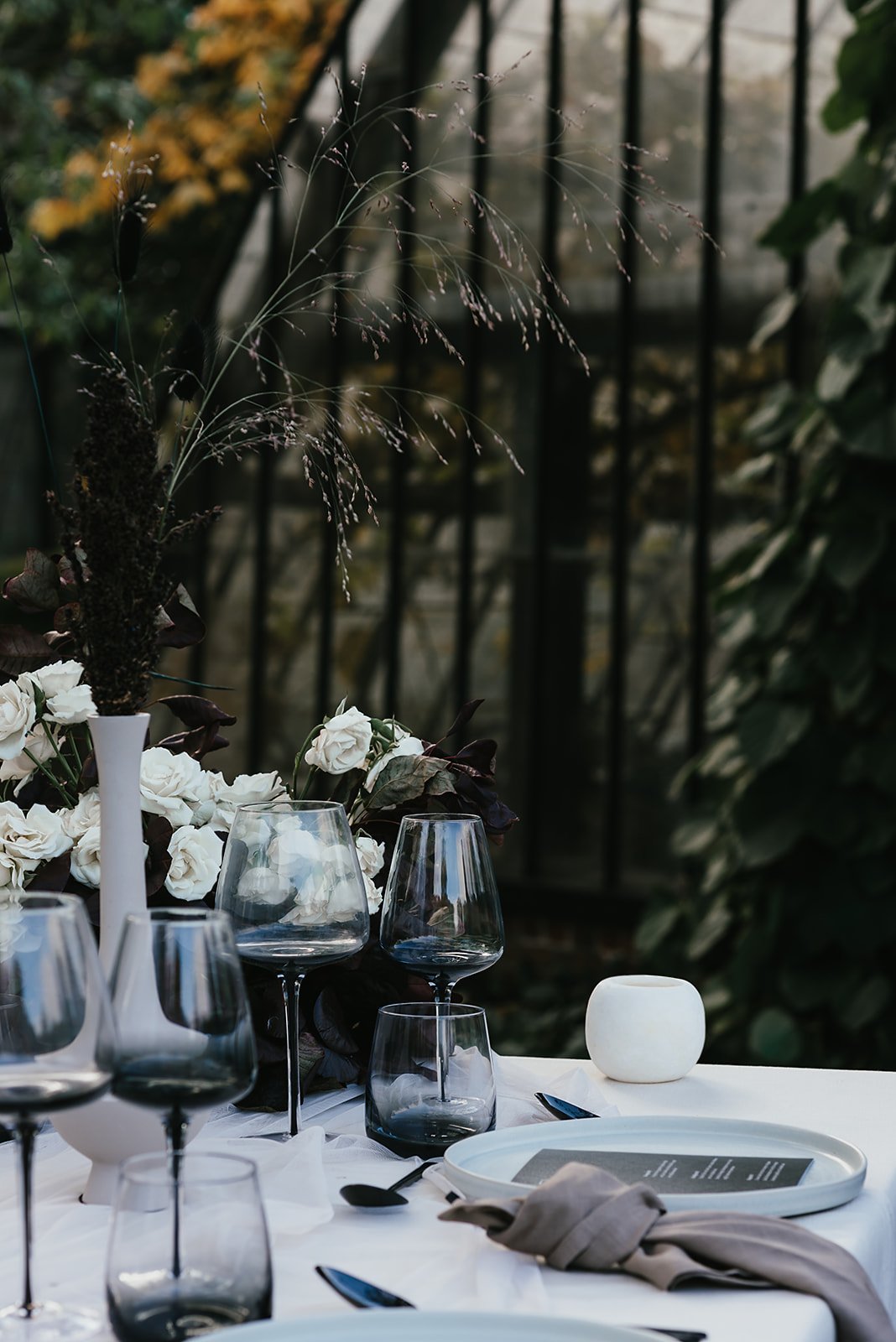 Elegant table setting with dark wine glasses, white plates, cloth napkin, candle, and floral arrangement in white vase outdoors.