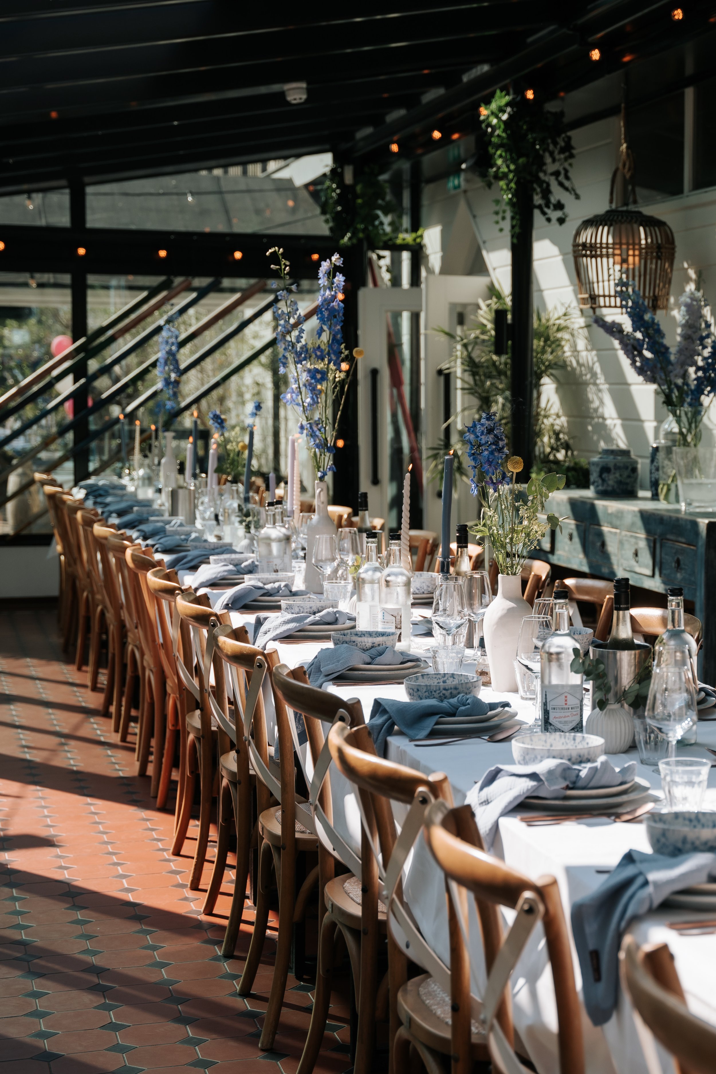 Lange, luxe gedekte eettafel voor een bruiloftsdiner. De ruimte is een glazen kas. Aan tafel staan houten crossback chairs. De tafelstyling is zomers blauw en wit, met blanco chique wijnglazen, whisky tumblers, verfijnde kaarsen en blauwe bloemen.