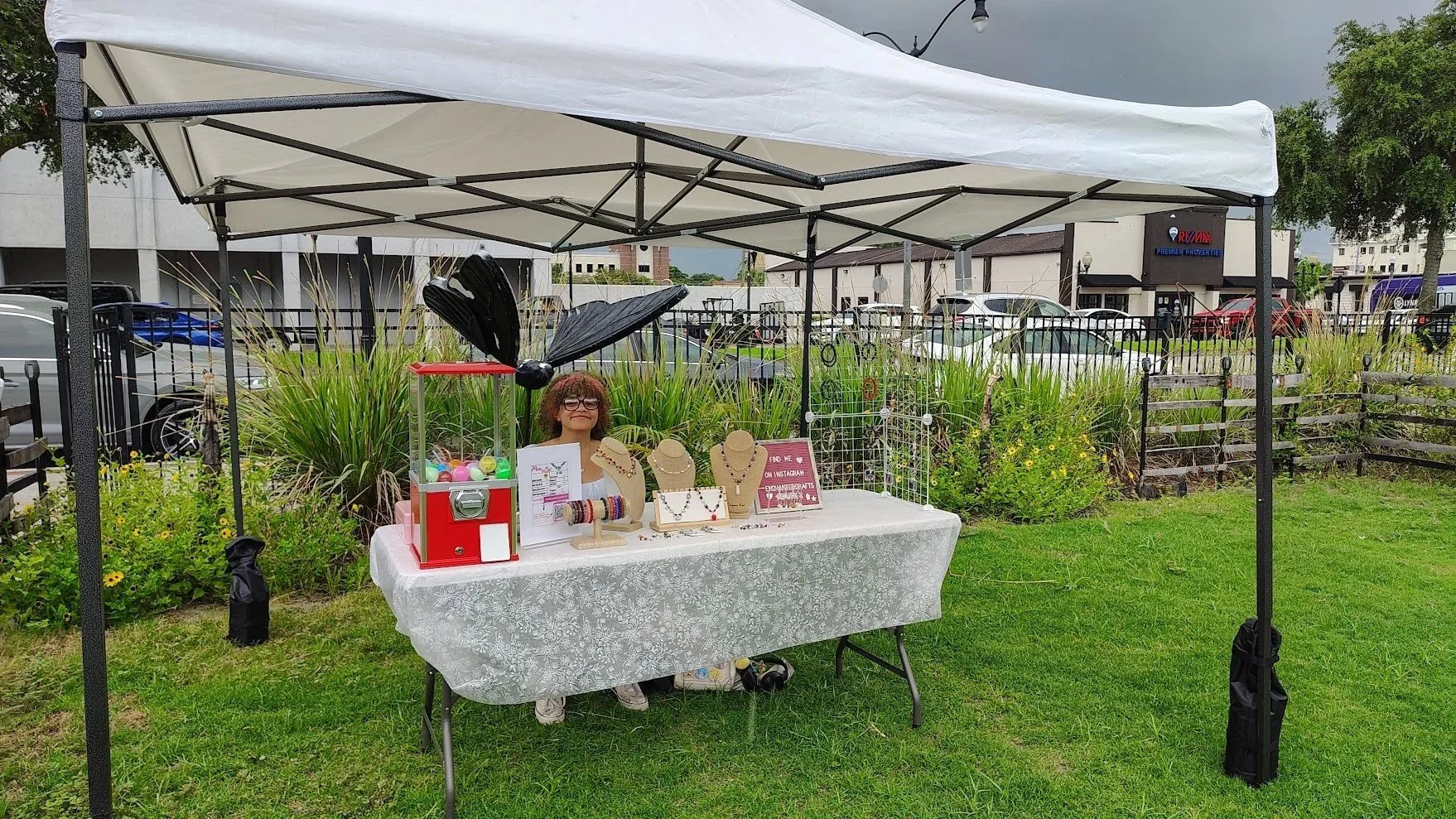 A woman displaying jewelry at an outdoor booth under a white canopy, with a gumball machine and various jewelry pieces on a table, surrounded by plants and parked cars in the background.