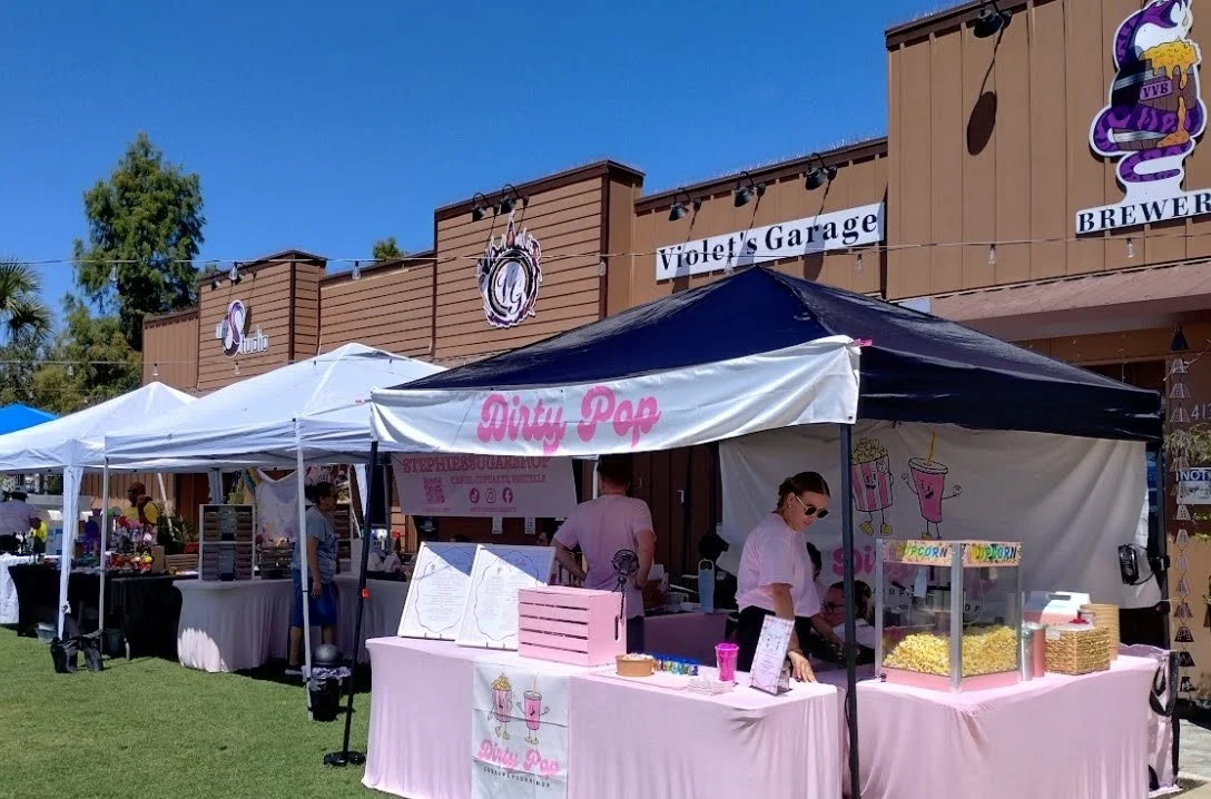 An outdoor market with various vendor tents, including one with a pink sign that reads "Dirty Pop" selling popcorn, in front of a building with signs that say "Violet's Garage" and "Brewery".