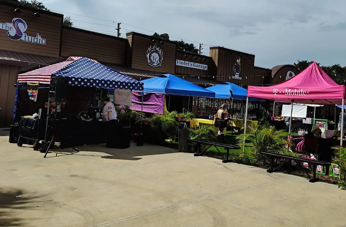 Outdoor shopping or event area with multiple vendor tents, including a pink T-Mobile tent, a blue and white star-patterned tent, and a blue tent, in front of a store building with signs for The Studio, Violet's Garage, and a penguin graphic.