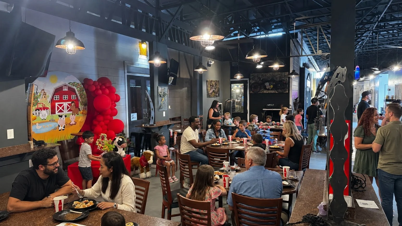 People dining in a restaurant decorated with farm-themed artwork and red balloon decorations, with children and adults enjoying meals, some standing and chatting near the entrance.