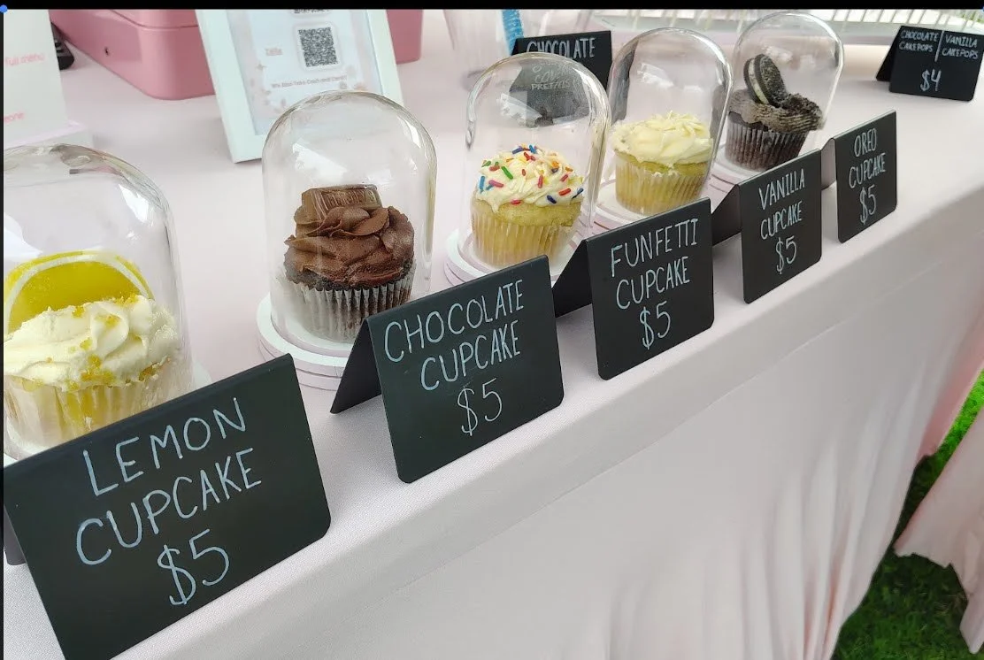 Display of cupcakes under glass domes with black labels showing flavor and price at an event stand at a Kissimmee Main Street market in Osceola County 