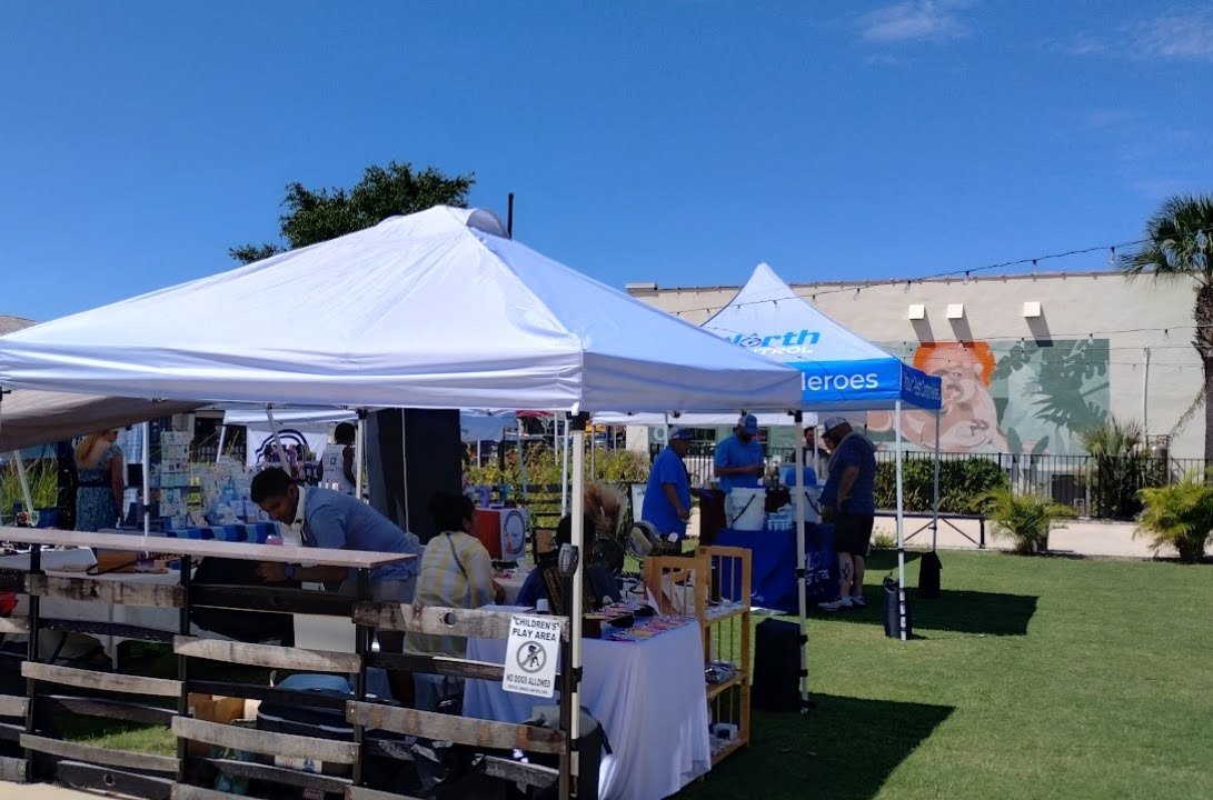 Outdoor market or event booths with white tents on a grassy area on a sunny day, with people browsing and socializing.
