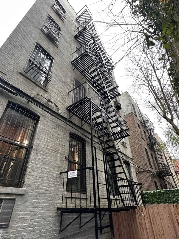 Black metal fire escape stairs and platforms climb a gray brick building with barred windows and exposed wiring.