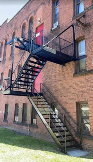 Two-level black fire escape with open metal stairs and railings connects to a red door on a brick building.