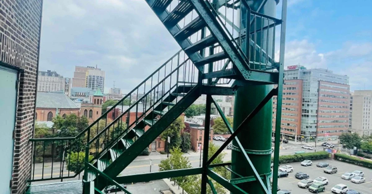 Green metal fire escape with multiple stair landings attached to a building, overlooking city streets and parked cars.