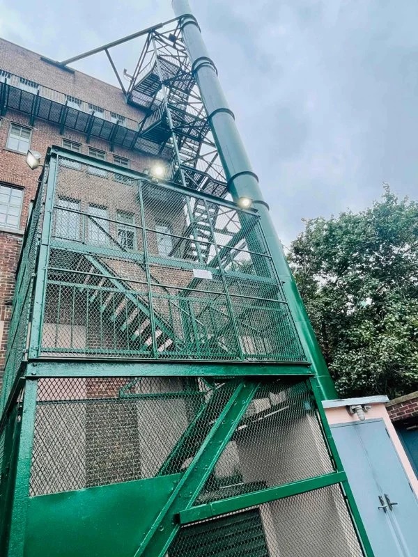 Green metal fire escape with mesh fencing and steep stairs rises beside a brick building and large vertical pipe.