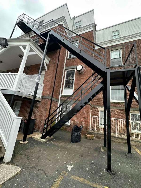 Black steel exterior fire escape with two stair flights attached to a multi-story red brick building beside a white porch.