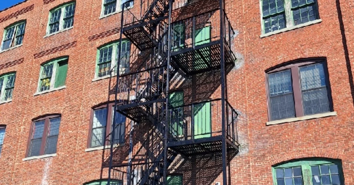 Black metal fire escapes attached to a red brick building with green window panels, many windows, and a blue sky.